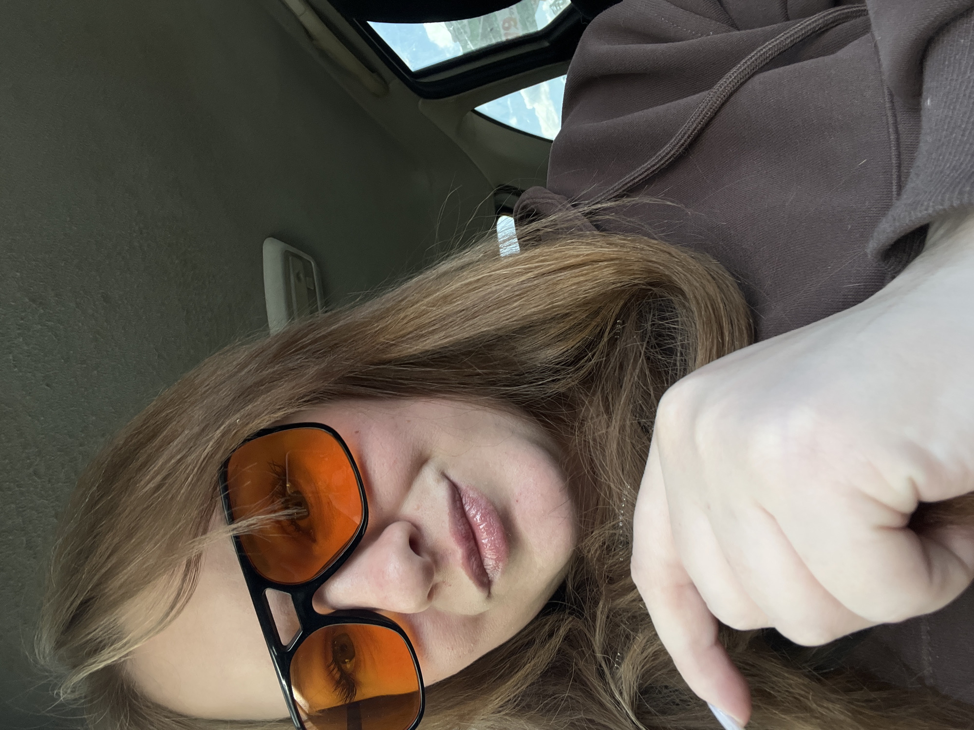 A selfie taken from a tilted perspective, possibly inside a car, shows a person with long, light brown hair and orange-tinted sunglasses. The individual is wearing a brown hooded sweatshirt. The background includes the car's interior, with the roof and a sunroof visible. The location of the photo is unknown, and there is no indication that it was taken in the city of Chaikovsky, Russia.
