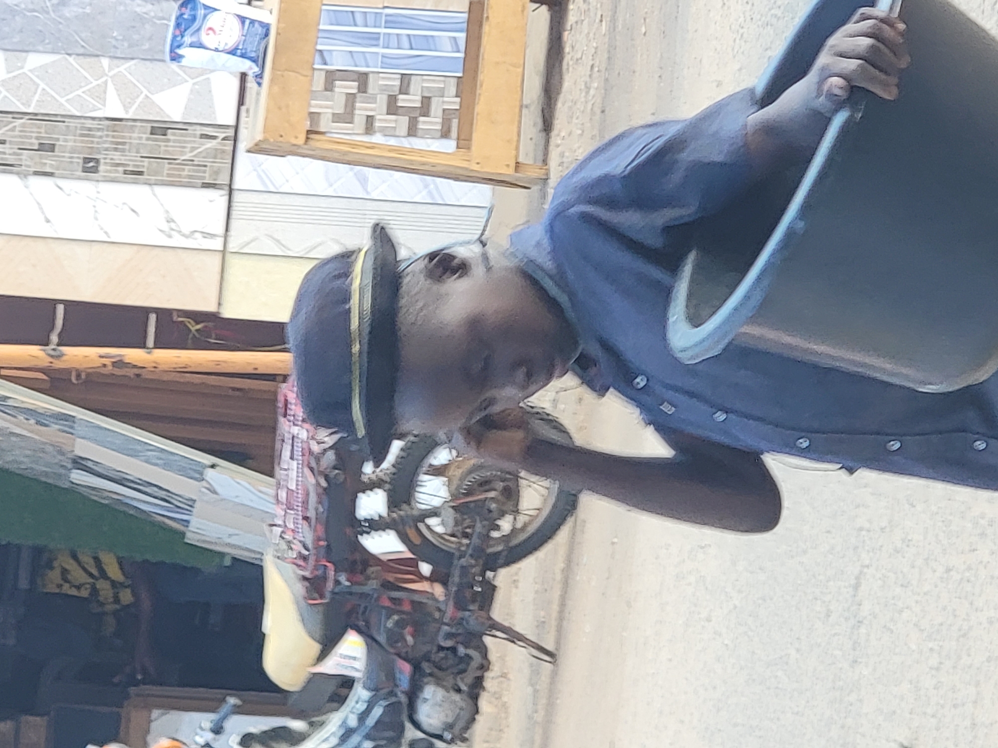 A person is carrying a black bucket in front of a shop in Maiburiji, Nigeria. The shop sells tiles, some of which are visible in their packaging. A motorcycle is parked nearby. The person is wearing a dark blue button-down shirt and a hat with a yellow band.