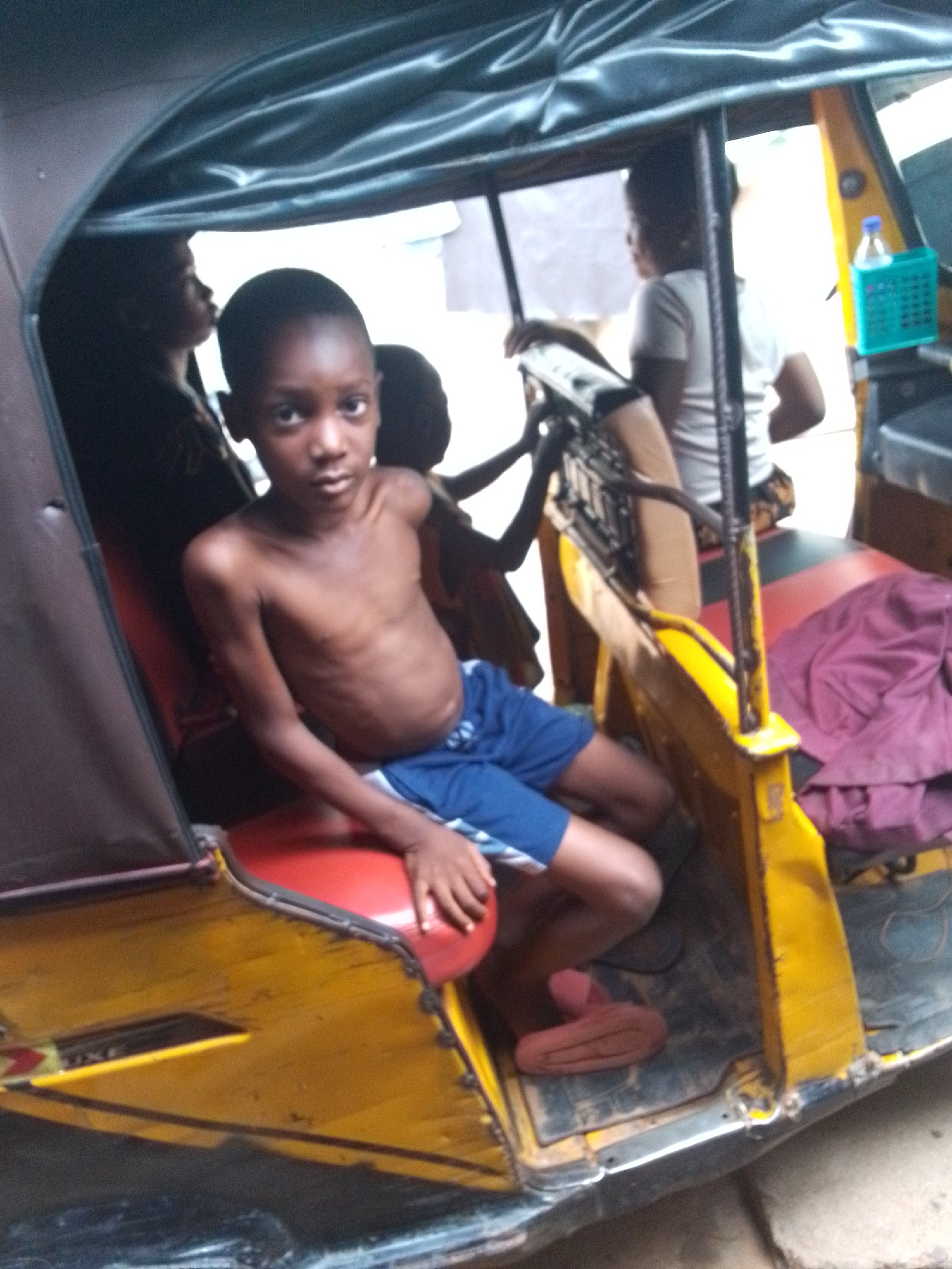 A medium shot captures a group of children inside a yellow motorized tricycle in Nkpor, Nigeria. The focal point is a shirtless boy in blue shorts, seated on a red seat and gazing directly at the camera. Other children are positioned behind him, one near the driver's seat and another visible in the background. A pink piece of fabric lies on the seat beside the main subject. The tricycle's interior is visible, and a blue container holding a water bottle is attached to the front panel. The environment suggests an outdoor setting.