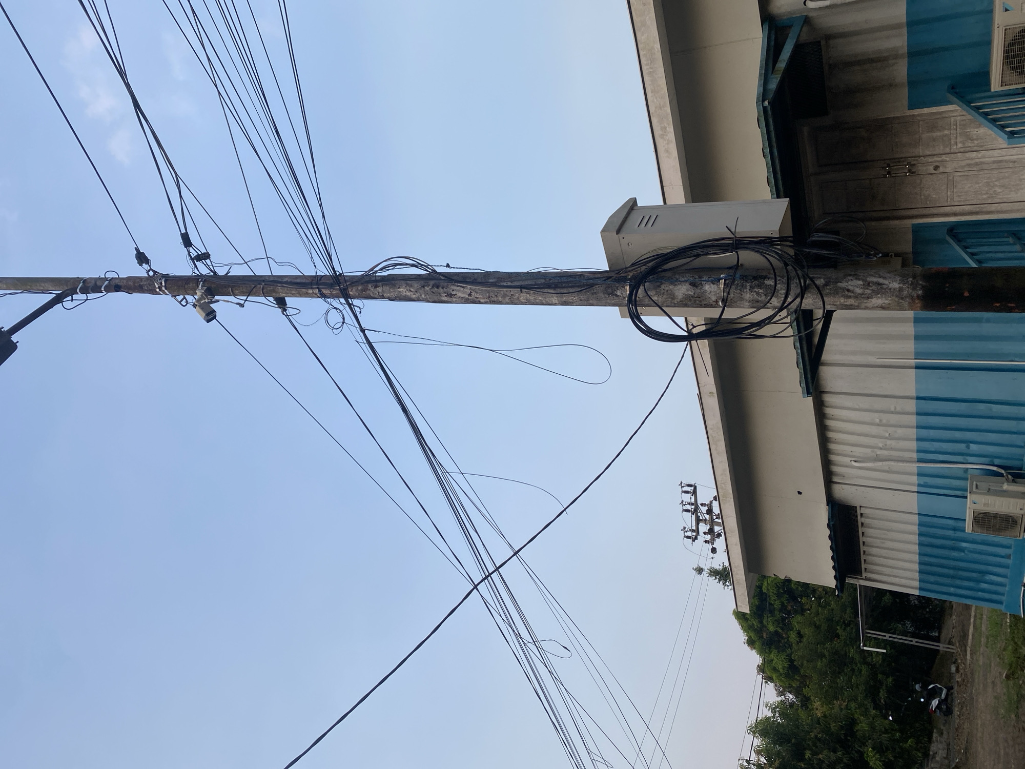 In Banjarbaru, Indonesia, a utility pole is the central focus of the image, situated in front of a building with a corrugated metal exterior painted in white and blue. The pole is adorned with numerous black electrical and communication wires, some of which are loosely coiled around it. A small, light-colored electrical box is mounted on the pole. A security camera is also visible attached to the pole. The building features a gray roof overhang above a set of wooden double doors and blue railings leading up to them. An air conditioning unit is installed near the base of the building. To the left of the building, there are trees and a parked white scooter. The sky above is a pale blue with a slight gradient, indicating it may be daytime or late afternoon.