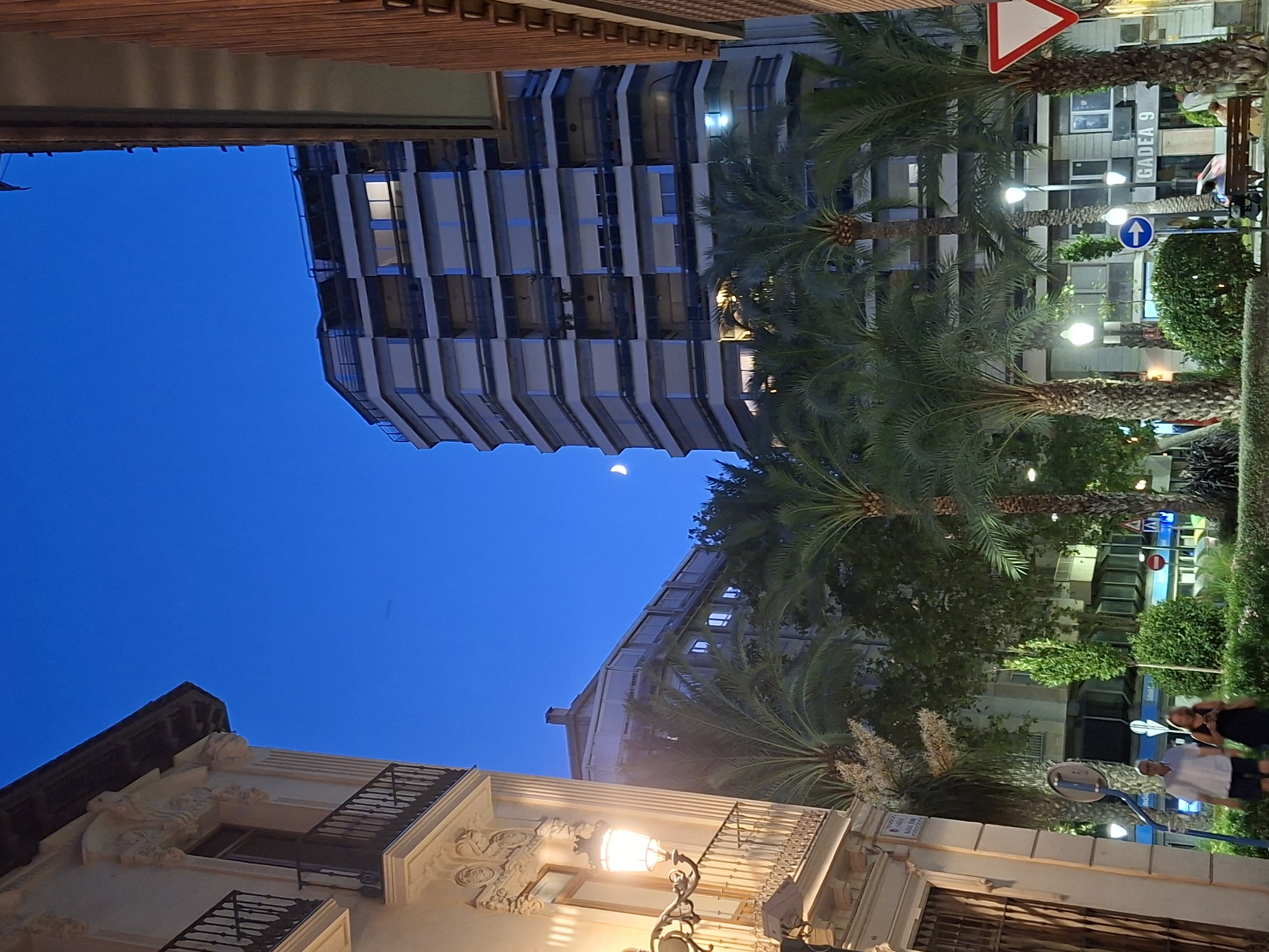 The image depicts an urban scene at dusk in Alicante, Spain. A tall, modern apartment building with numerous balconies dominates the right side of the frame, its windows illuminated. In the foreground, palm trees are visible, suggesting a Mediterranean climate. To the left, an older building with ornate architectural details and a balcony is partially visible. A streetlamp on this building is lit. A crescent moon is faintly visible in the deep blue twilight sky. Two people, a man and a woman, are walking away from the camera on a sidewalk. The man is wearing a white shirt and dark shorts, while the woman is wearing a dark dress. A street sign with a blue arrow is present, and another sign reads "GADEA 9". The overall impression is that of a city street at the end of the day.