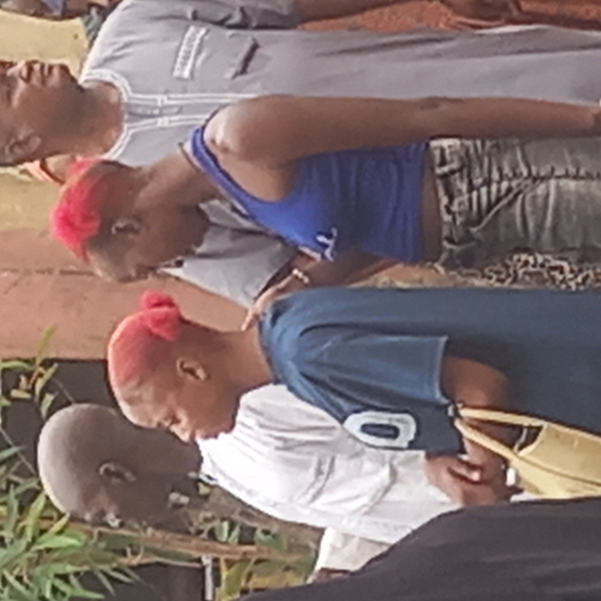 A medium, color shot captures several people, including two women with red hair, bending their heads downward. The women are wearing blue tops. To the right of the second woman is a bald man wearing a white shirt. The people are standing outside in what appears to be Dadi Riba, Nigeria.