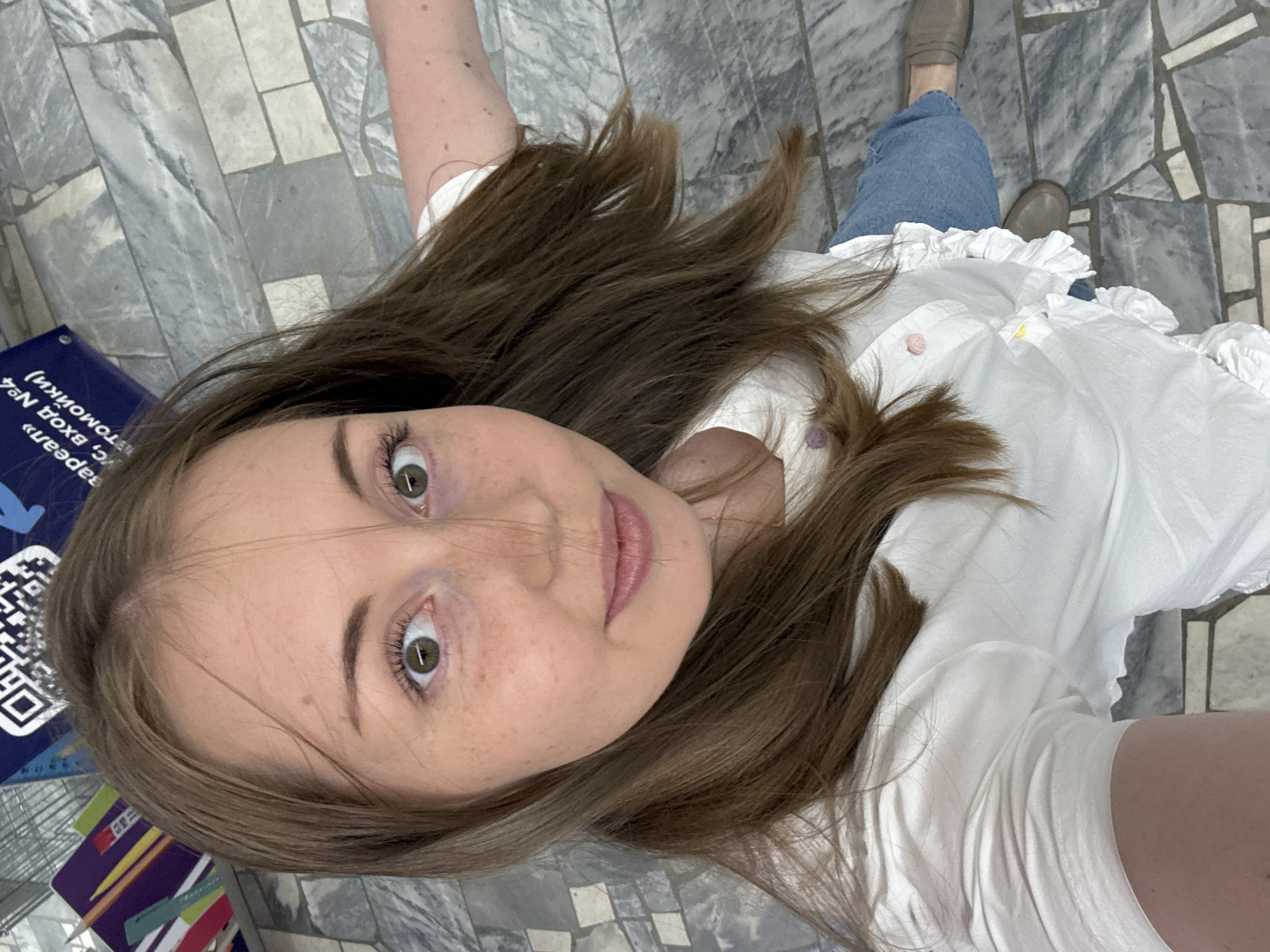 A woman is taking a selfie from a high angle. She has medium-length brown hair, green eyes, and light skin with freckles. She is wearing a white ruffled blouse and blue jeans. The background is a tiled floor and a blue sign with text and a QR code. The woman's arm is extended to the right, suggesting she is holding the camera or phone. Her expression is neutral.
