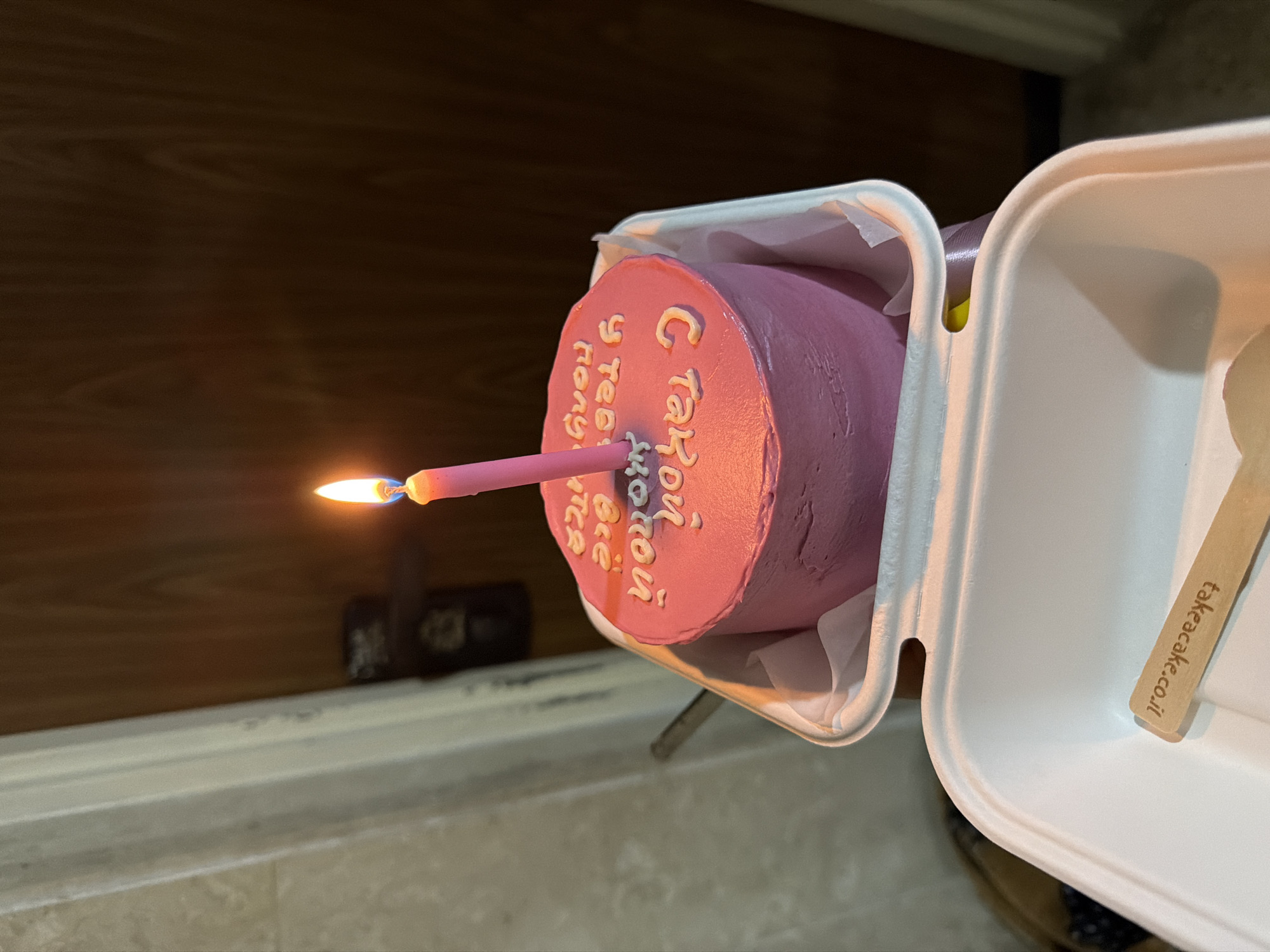 A tilted overhead shot reveals a small, round cake with pink frosting, decorated with white lettering in Cyrillic reading, "С Такой чудной веселой и Тебя с днем рожденья". A single, pink birthday candle stands upright in the cake, its flame flickering. The cake is presented in a white, disposable container next to a wooden utensil branded with "takeacake.co.il", indicating the cake’s origin is likely in Israel. The setting appears to be indoors, with a dark wooden wall visible in the background. While the image location isn't stated, the domain "takeacake.co.il" suggests the city is Rishon LeZion, Israel. There are no people visible in the scene.