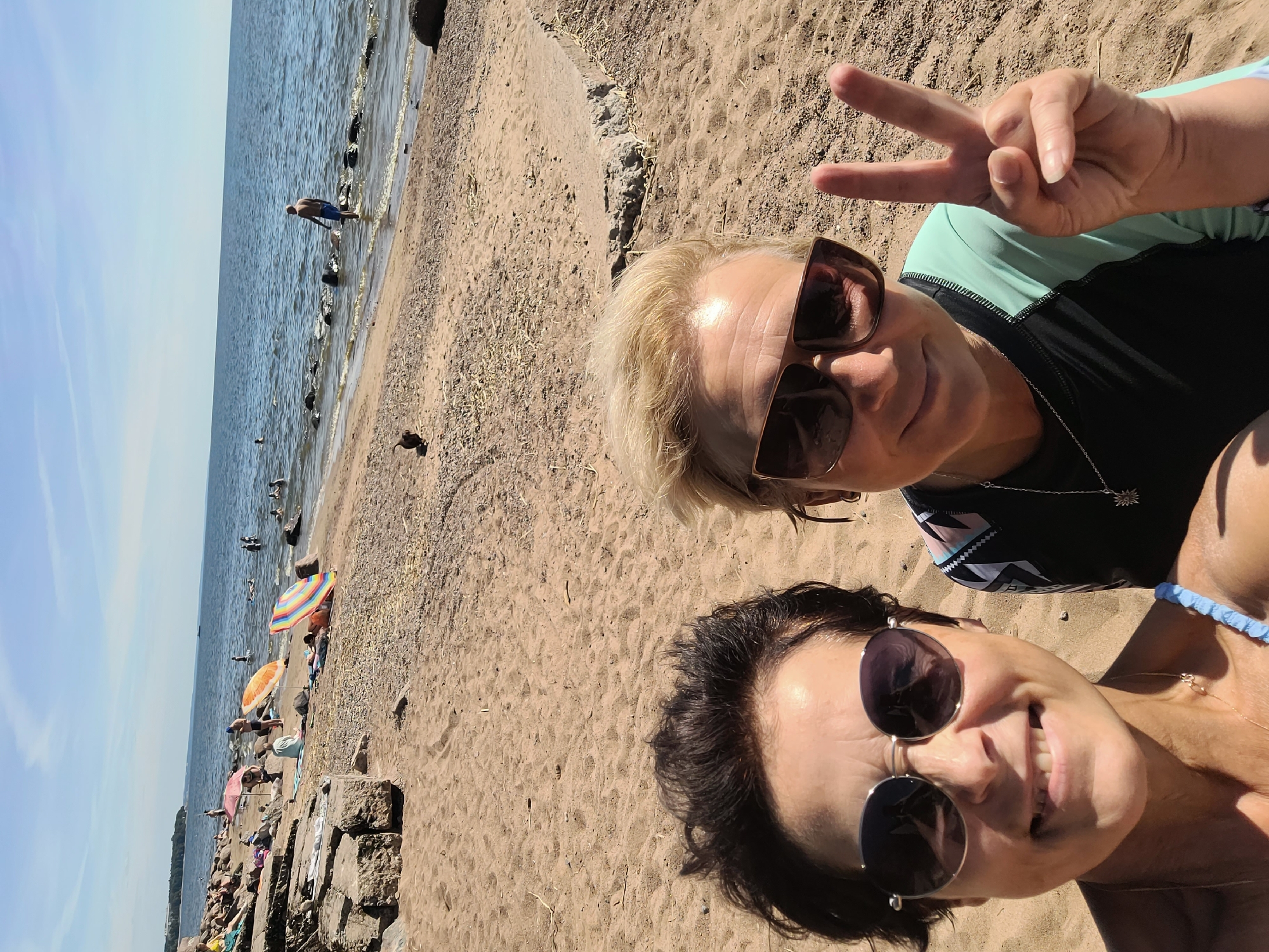 Two women are posing for a selfie on a sandy beach in Repino, Russia. The woman on the left has short, dark hair and is wearing round sunglasses. The woman on the right has blonde hair and is wearing oversized sunglasses. She is holding up two fingers in a peace sign. In the background, people are swimming in the sea, relaxing on the beach under umbrellas, and walking along the shore. The water is calm and the sky is blue with some scattered clouds. The beach is a mix of sand and pebbles, with large rocks visible along the coastline.