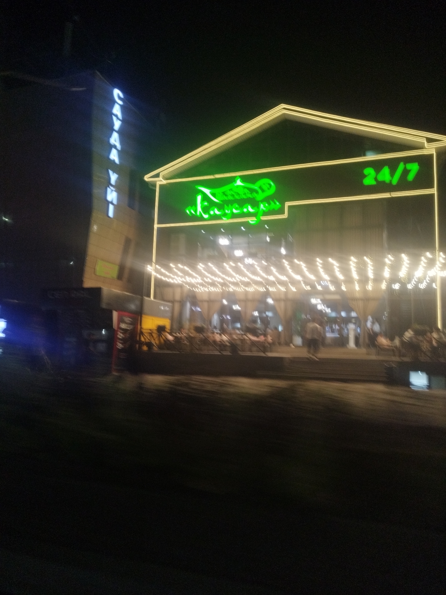 The image shows a nighttime street scene in Shymkent, Kazakhstan. A neon sign for "Sayahaт" is visible vertically on the side of a building. To the right, a building with a stylized roof outline is illuminated by a neon sign that reads "24/7" and appears to have a graphic of a fish. String lights are draped across the front of the building, illuminating an outdoor seating area where several people are visible, seated at tables or standing. The overall impression is that of a restaurant or entertainment venue.
