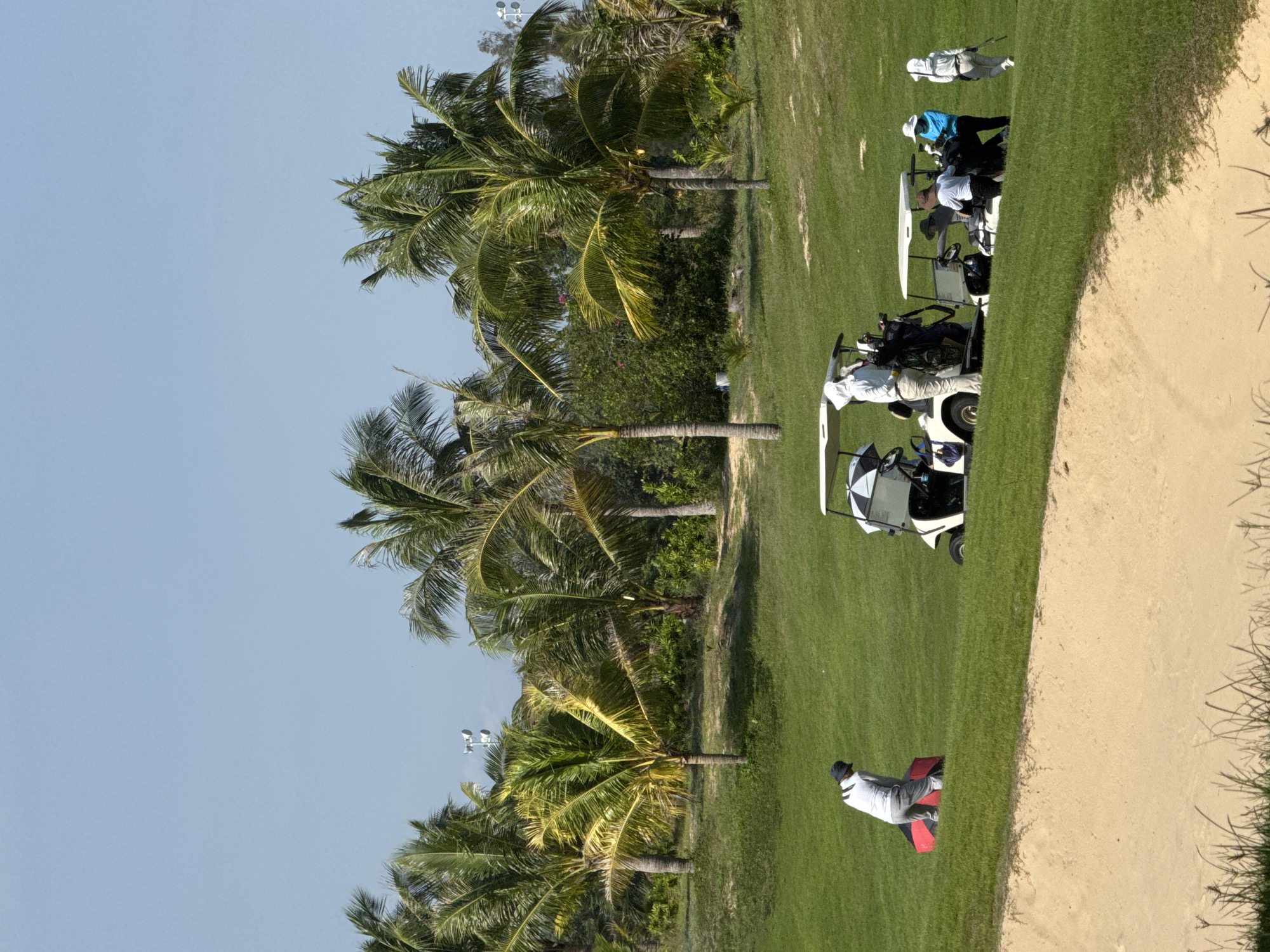 The image depicts a golf course scene in Phú Thủy, Vietnam. Several people are present, engaged in golfing activities. In the foreground, a sandy bunker is visible. Further back, on the green grass, a person in a white shirt and grey pants is preparing to hit a golf ball, with a red bag beside them. Two golf carts are parked on the course. One cart, on the left, is partially obscured by a person standing near it. The second cart, further to the right, has several people gathered around it. One person in a blue shirt is interacting with the cart, while others in white attire stand nearby. In the background, lush green palm trees and other vegetation create a tropical environment. The sky is clear and blue.