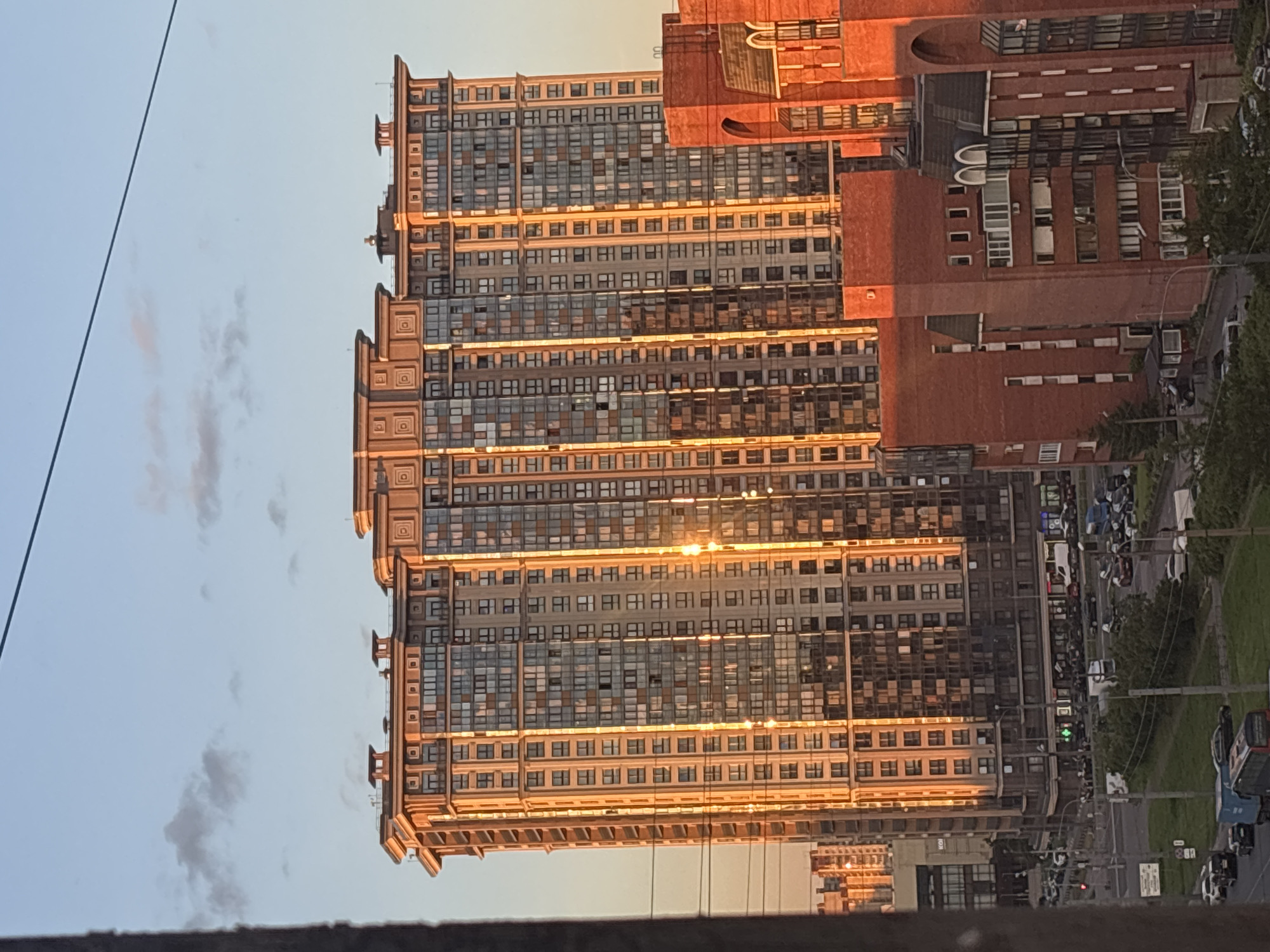 A golden hour shot captures a tall, modern apartment building in an urban setting. The building's facade is a mix of beige and glass, with numerous windows reflecting the warm sunlight. The structure features a decorative top with multiple rectangular elements. In front of the taller building, a shorter, reddish-brick building is visible, with arched window details on its right side. The lower portion of the image shows a street scene with various vehicles, including cars and trucks, and some greenery. The sky above is a light blue with scattered clouds. The image appears to be taken from a higher vantage point, possibly another building.