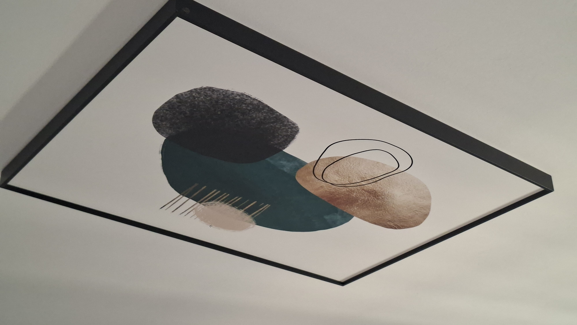 This is a medium shot, eye-level, indoor photograph of a framed abstract art piece hanging on a white wall. The frame is thin and black, and it is angled slightly to the right. The artwork itself features several abstract shapes in muted colors, including a dark teal oval, a mottled dark gray shape, a light pink oval with thin gold lines extending from it, and a metallic copper oval with black line drawings of a circle and an oval superimposed on it. The lighting appears to be ambient, casting soft shadows on the wall behind the artwork. There are no people or any other discernible objects in the scene. The image does not provide any information that would allow for its location to be identified in Podgorica, Montenegro.