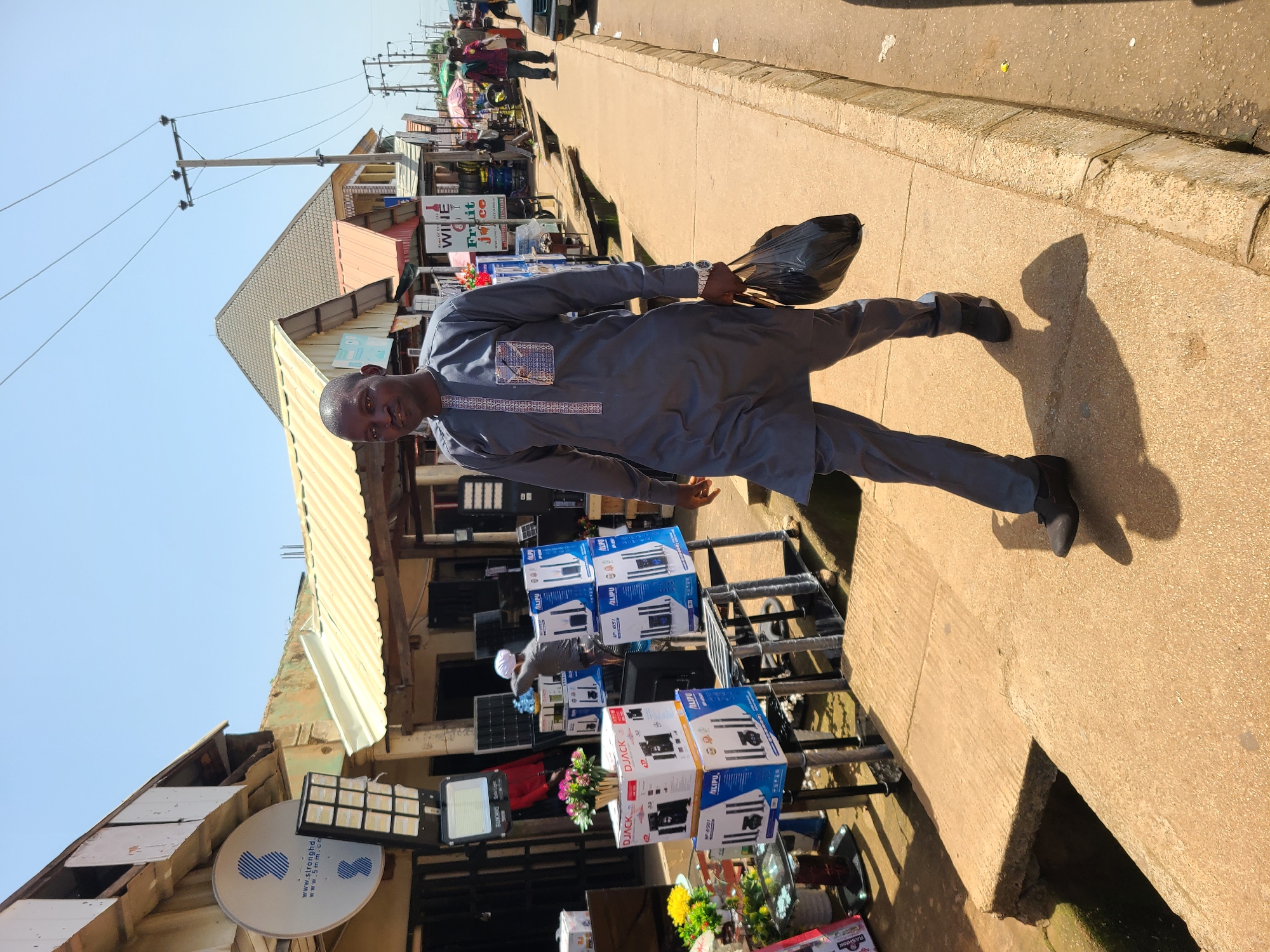 In Bukuru, Nigeria, a man walks along a concrete sidewalk next to a street lined with shops and market stalls. He is wearing a grey, long-sleeved tunic and trousers, and carrying a black plastic bag in his right hand. The man is walking towards the right of the frame, with his body angled slightly towards the camera.
The street is a mix of paved and unpaved surfaces, with drainage channels visible along the side. Shops line the sidewalk, displaying a variety of goods, including electronics, home appliances, and packaged drinks. Some stalls are covered by awnings or temporary structures. The sky is clear and bright, indicating a sunny day. In the background, more shops and people are visible, as well as power lines and utility poles. The scene depicts a bustling urban environment with commercial activity.