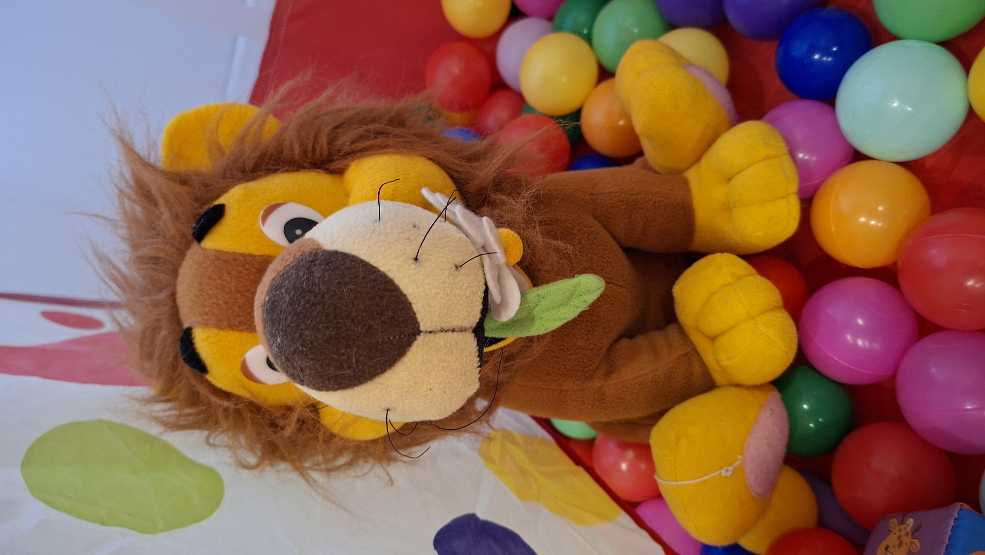 A stuffed lion with a brown mane and yellow and tan face lies on a red, white, and yellow patterned surface. The lion is partially surrounded by an assortment of colorful plastic balls.
