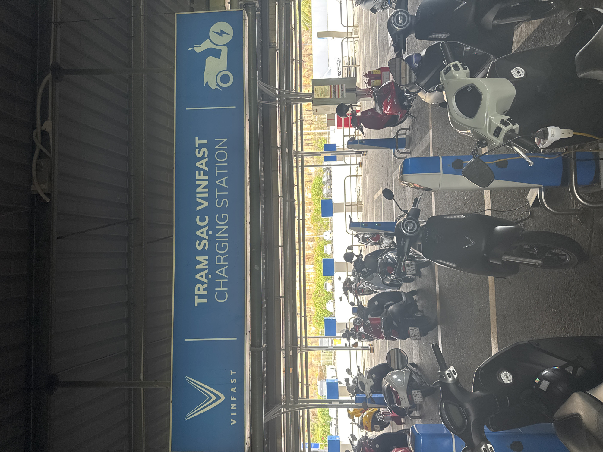 The image shows a VinFast charging station for electric scooters, likely located in Đà Nẵng, Vietnam. A sign with the VinFast logo and the words "TRẠM SẠC VINFAST CHARGING STATION" is prominently displayed. The station is under a covered structure, and multiple scooters are parked in charging bays. Some scooters are actively being charged, indicated by the charging cables connected to them. The scooters are varied in color, including red, black, and white. License plates are visible on some scooters. The environment appears to be a parking facility with other vehicles visible in the background.