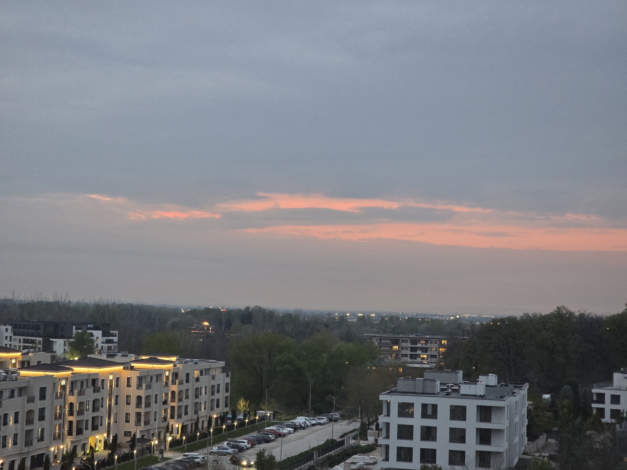 The image presents an elevated view of a residential area at dusk or dawn. The sky is largely overcast with soft grey clouds, but a prominent band of delicate pink and orange clouds stretches across the horizon, indicating either a recent sunset or an impending sunrise.
In the foreground and mid-ground, several modern residential buildings are visible. To the left, a large, multi-story building with a light-colored facade features numerous balconies, arched windows, and intricate architectural details. This building is brightly illuminated with warm artificial lights, including strip lighting under its cornices and facade uplights, suggesting a contemporary design. Parked cars line the street adjacent to this building.
To the right, another distinct, modern white building with square windows and balconies stands out. Further in the mid-ground, a dense expanse of trees, appearing to be a forest or a large park, separates different parts of the urban landscape. Beyond the tree line, more distant buildings and scattered lights can be seen, hinting at a wider urban sprawl. Streetlights are illuminated along the roads and pathways, and some lights are visible inside the residential units. No human activity is overtly depicted, and no discernible text is visible within the frame. The overall atmosphere is quiet and serene.