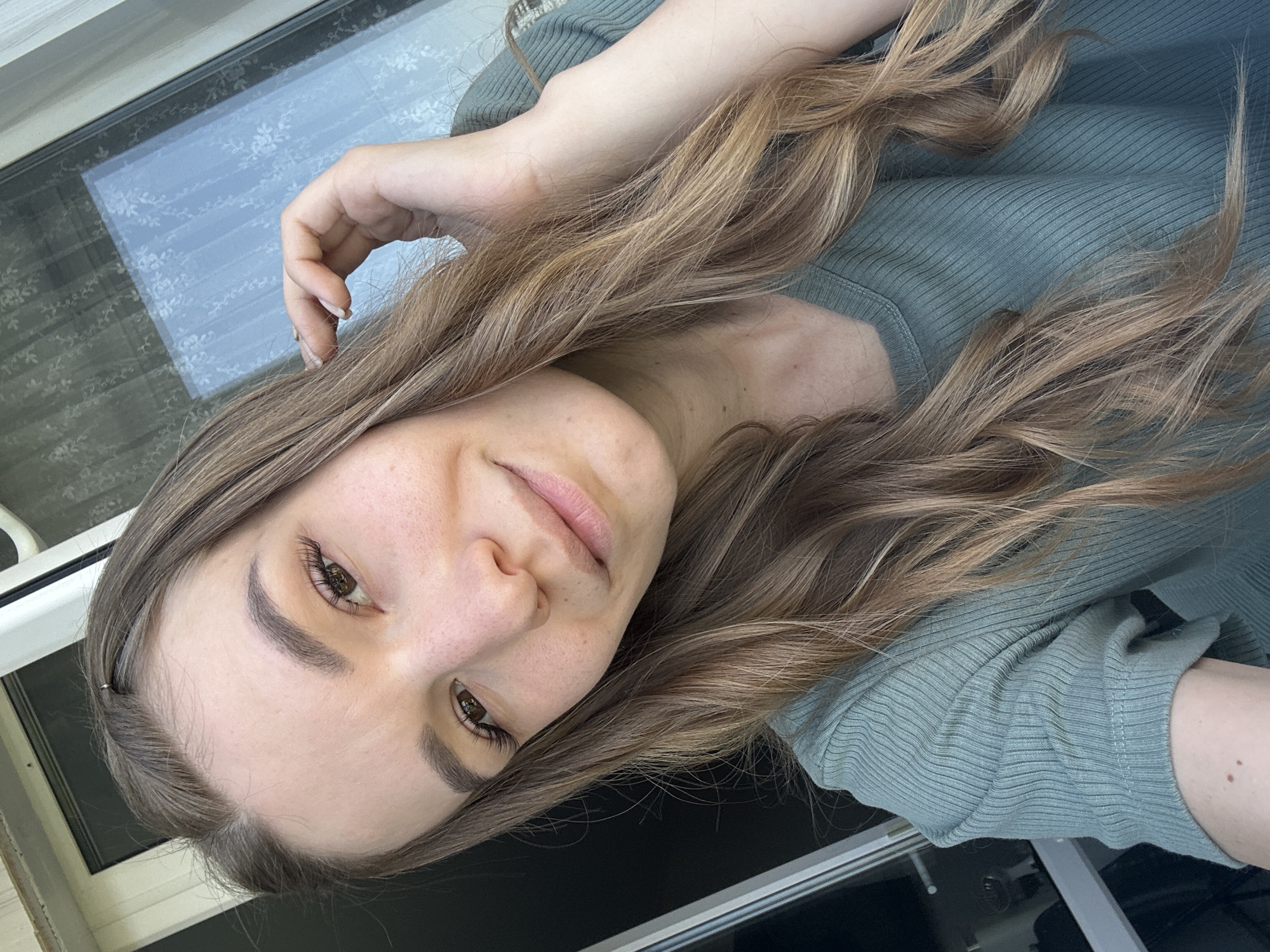 A woman is posing for a selfie indoors. The photo is rotated counter-clockwise so that her left side is at the top of the frame. She has long, light brown hair and is wearing a light green ribbed shirt. Her left hand is raised and resting on her hair. A window is visible in the background. It cannot be definitively determined if the image was taken in Chaikovsky, Russia.