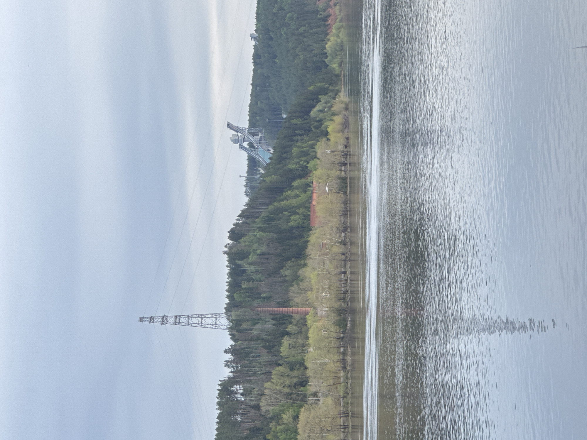 The image depicts a landscape in the city of Chaikovsky, Russia. A body of water occupies the right side of the frame. The opposite bank is lined with trees. A power line tower is visible in the lower left of the frame. Further up the bank is a brick structure, and a ski jump. The sky is overcast.