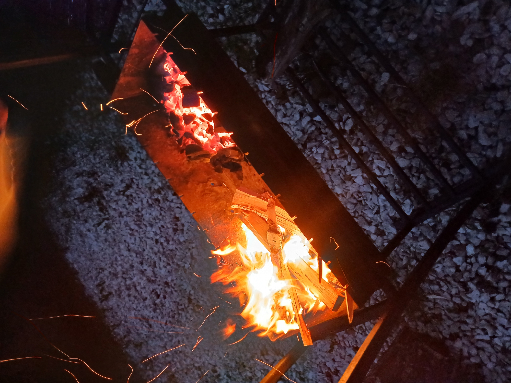 In Verkhneye Dubrovo, Russia, a close-up, high-angle shot captures the fiery heart of a campfire. The scene is dominated by a metal fire pit filled with glowing embers and burning logs. Flames, rendered in vivid oranges and yellows, dance upwards, illuminating the surrounding wood. Tiny, bright orange sparks are scattered across the frame, suggesting the active nature of the fire. The fire pit is surrounded by gravel, which appears somewhat damp, and a dark, metallic grill structure is visible to the right of the pit. The overall lighting suggests it is nighttime or dusk, with the fire providing the primary light source. There are no people or animals visible in the frame, and no text is present. The mood is one of warmth and natural energy, highlighting the raw power of the fire in a rustic outdoor setting.