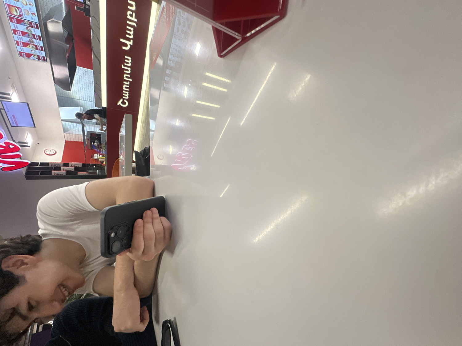The image captures a scene in what appears to be a fast-food restaurant or food court, with the camera oriented sideways. In the foreground, a person with dark, curly hair and wearing a white t-shirt is leaning on a white, reflective table. They are smiling and looking down at a dark-cased smartphone, which they hold horizontally with both hands. Another person is partially visible in the bottom left, wearing a dark top.
The background reveals the interior of the establishment. To the right of the visible person, a tall red sign with white Armenian text is prominent. The text reads "Դառնամ Վայթը" (Darnam Vayty), and above it, in a smaller font, "Արագ" (Arag, meaning Fast). Further back, there's a serving counter with menu screens displaying food items, and another individual can be seen standing behind the counter. A large, partially cropped red logo, resembling a stylized "K" and "C" (similar to the KFC logo), is visible on the far left side of the background. The entire area is brightly lit by overhead strip lights, which reflect on the shiny table surface. The person's smile suggests a relaxed and positive mood.