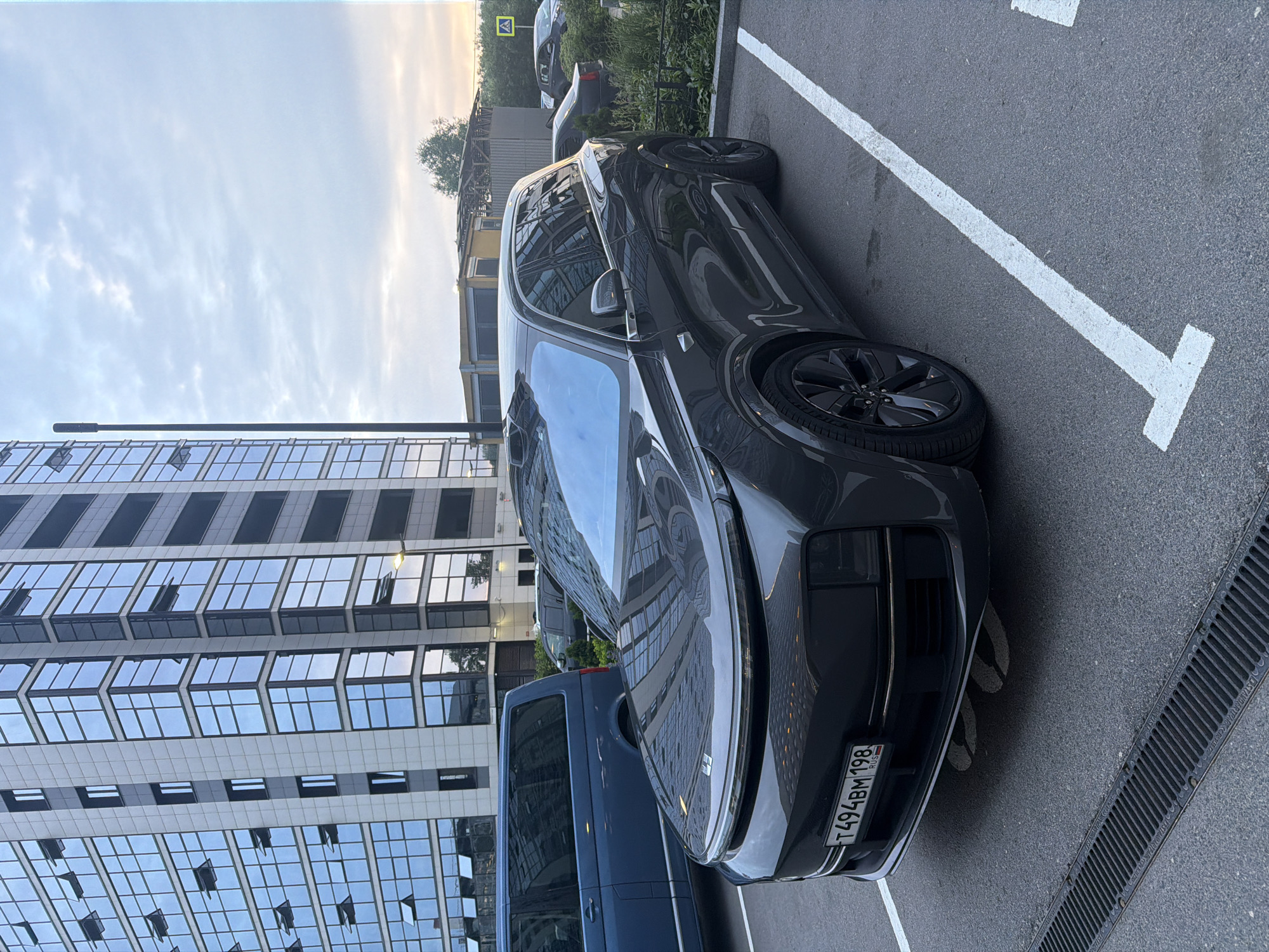 A dark gray SUV is parked in a parking lot in front of a modern, multi-story building with a glass facade. The SUV has a license plate that reads "T494BM 198 RUS". To the left of the SUV, a dark blue minivan is parked. In the background, there are other parked cars and some greenery. The sky is overcast, and it appears to be either early morning or late evening. The scene is in a city, but the specific location is unknown.