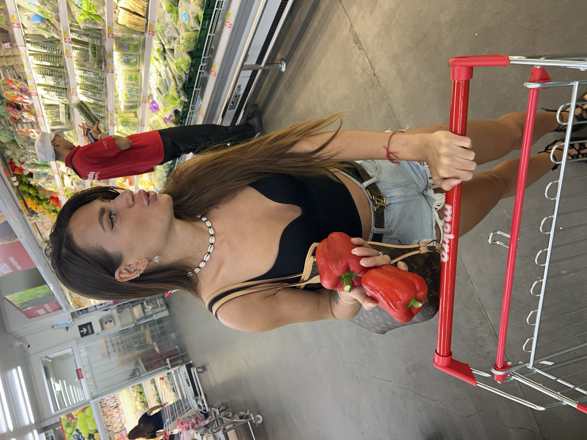 A woman with long brown hair is standing in a grocery store, holding two red bell peppers and a shopping cart. She is wearing a black crop top, denim shorts, and a necklace made of shells. Her left hand is holding the peppers, and her right hand is on the shopping cart. In the background, a man in a red shirt and a white hat is looking at some produce, and other shoppers and displays are visible. The floor is gray concrete, and the shelves are stocked with various fruits and vegetables. The setting appears to be a modern supermarket.