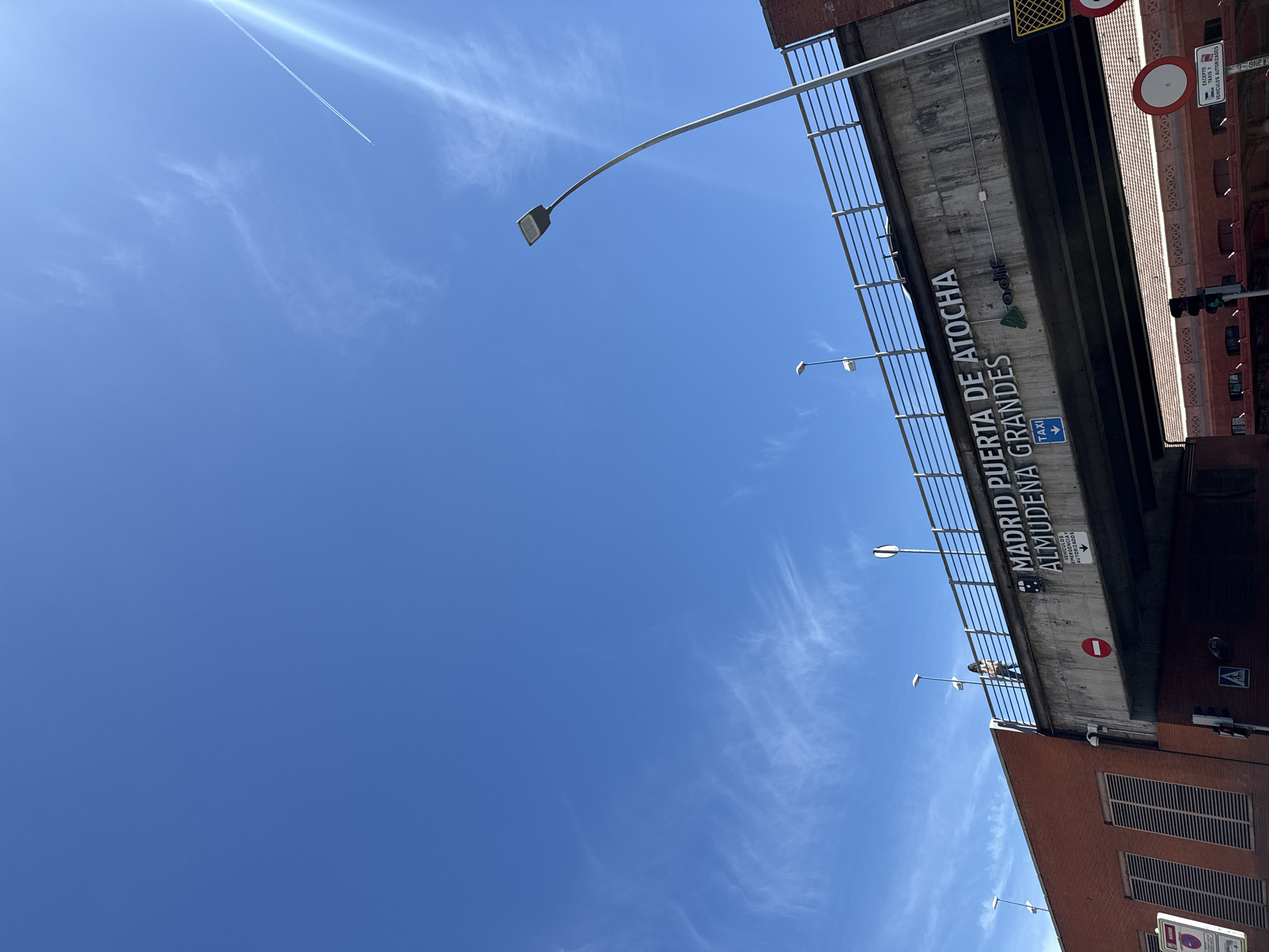 This media file, originally rotated 90 degrees counter-clockwise, depicts a bright, sunny daytime scene in Madrid, Spain. The vast upper portion of the correctly oriented image shows a clear, vibrant blue sky, sparsely dotted with wispy white clouds. A prominent, luminous contrail from an airplane stretches diagonally across the upper left corner of the sky.
The lower half of the image is dominated by an urban overpass or bridge made of concrete, equipped with a metal railing. Attached to the side of the overpass is a large white sign against a dark background, clearly readable as "MADRID PUERTA DE ATOCHA ALMUDENA GRANDES," with the "adif" logo also visible, indicating a location near the Atocha train station. Below this, a blue sign with a downward arrow points to "TAXI." Other visible road signs include a red circular "no entry" sign and a square sign that states "EXCEPTO TAXIS VEHÍCULOS AUTORIZADOS" (Except taxis authorized vehicles), suggesting controlled access to the area.
Multiple streetlights with modern, rectangular lamps are mounted on the overpass and in the foreground. One person is visible on the overpass, leaning against the railing. To the right, part of a brick building with dark, slatted windows is visible, typical of an urban setting. The time of day is clearly daytime, under a bright sun with fair weather.
