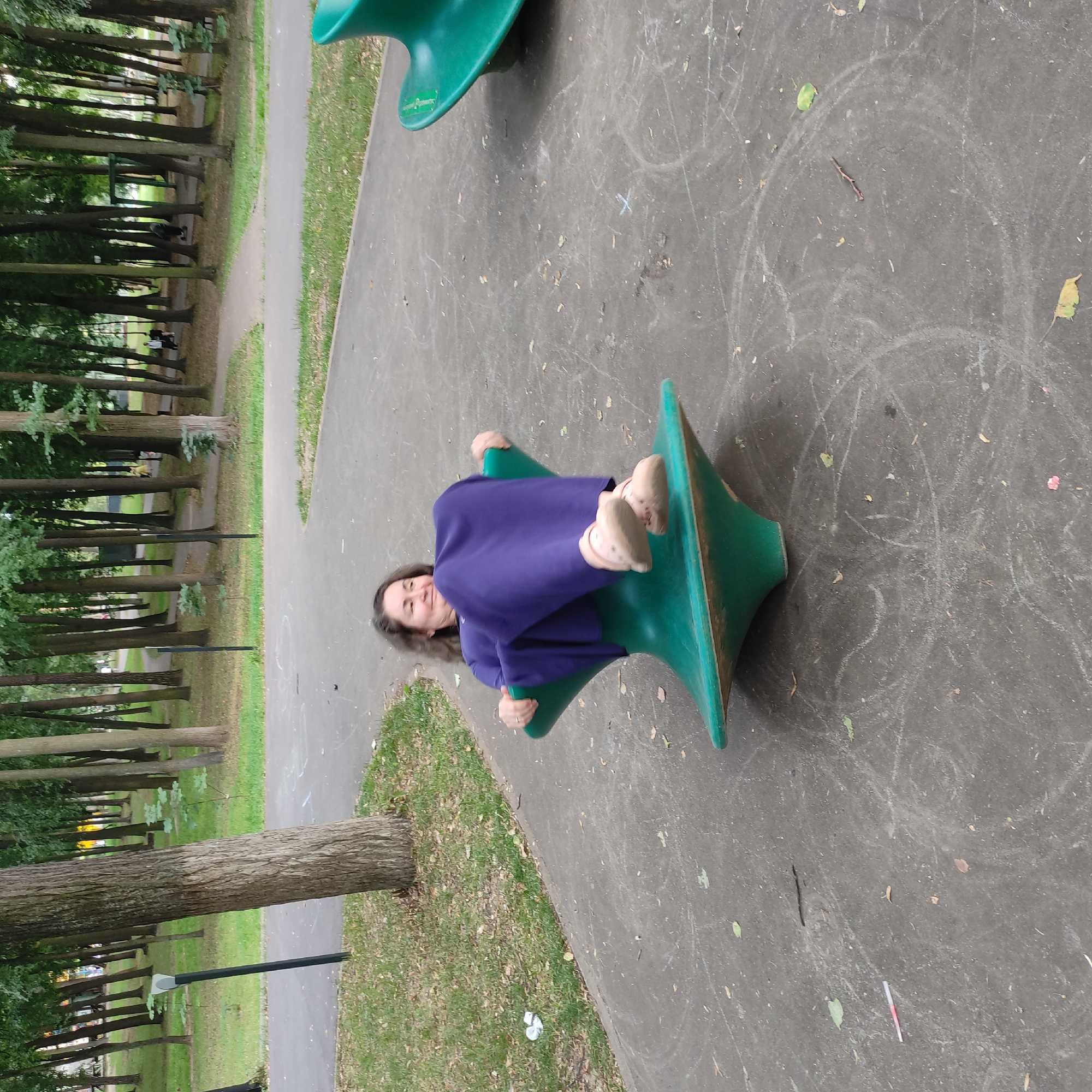 In a park setting, a woman is seated on a green spinning playground toy, which resembles a large spinning top. She is wearing a long, flowing purple dress and light-colored shoes. Her hands are gripping the sides of the toy as she leans back with a smile, her legs drawn up. The toy is positioned on a paved surface, likely a playground or park path, with chalk marks visible on the ground. Surrounding the paved area is grass and a dense collection of trees, suggesting a wooded park. The scene is captured from a slightly elevated perspective, looking down at the woman and the toy. The background shows more trees and a pathway winding through them.