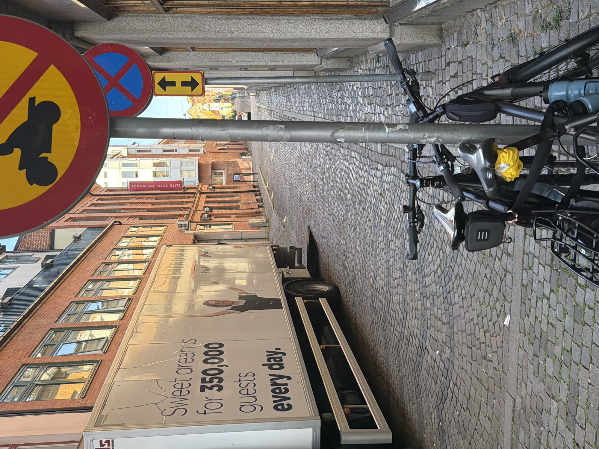 The image depicts a cobblestone street in Malmö, Sweden, likely in the Gamla Väster area, characterized by brick buildings with multiple windows and a truck parked along the left side. The truck has a large advertisement on its side, with text that appears to read "Sweet dreams for 350,000 guests every day." A woman in a dark uniform is pictured on the truck.
Several traffic signs are visible, including a "no motorcycles" sign, a "no parking" sign (blue circle with a red cross), and a directional sign with up and down arrows. Two bicycles are parked near a lamppost in the foreground. The buildings on the right side of the street have stone facades and wooden window frames. The street is narrow and appears to be in a pedestrian-friendly or historically preserved district. The lighting suggests it is daytime, possibly late afternoon given the shadows.
