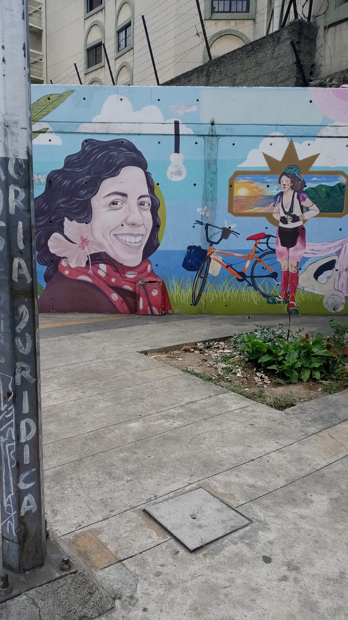 The image shows a concrete sidewalk in front of a mural on a wall. The mural features a portrait of a woman with dark curly hair and a red scarf, a bicycle, a woman with a crown wearing a backpack and boots, and a painting depicting a sunset over a landscape. The mural also includes elements like clouds and a guitar. A metal pole with graffiti is situated in the foreground. A small patch of greenery is visible to the right of the sidewalk, and there is a building in the background with windows and barbed wire on top of the wall. The city and exact location are unknown.