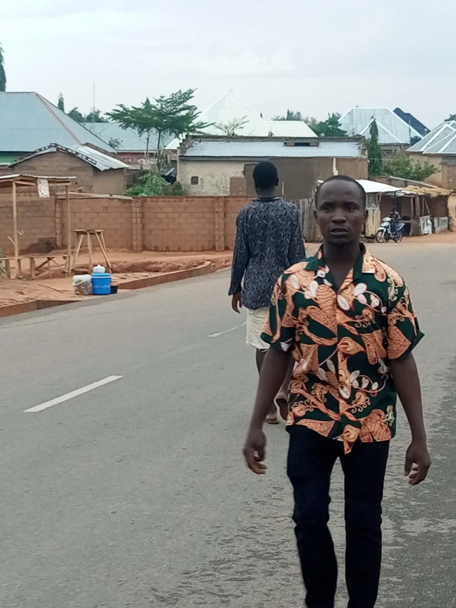 The image shows two men walking on a street in what appears to be a residential area. The foreground figure is wearing a patterned shirt and dark pants, facing the camera. Behind him, another man is walking away, wearing a darker shirt and light-colored shorts. In the background, there are buildings, some with corrugated metal roofs, and a motorcycle with a rider. There is a low brick wall and a roadside setup with water containers. The scene is set on a paved road with a white line marking. The location is Tempure, Nigeria.