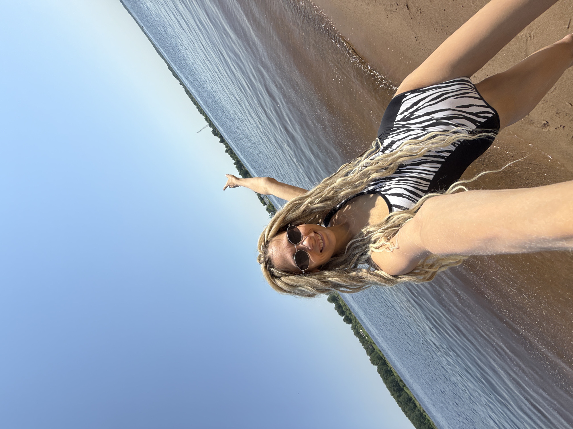 A young adult female is pictured taking a selfie at a beach in Rusanovo, Russia. She is wearing a zebra-striped swimsuit and sunglasses. Her blond hair is styled in braids. She is smiling and pointing with her left hand towards the horizon. The background consists of a beach, water, sky, and a distant treeline. The water is gently lapping at the shore. The sky is blue.