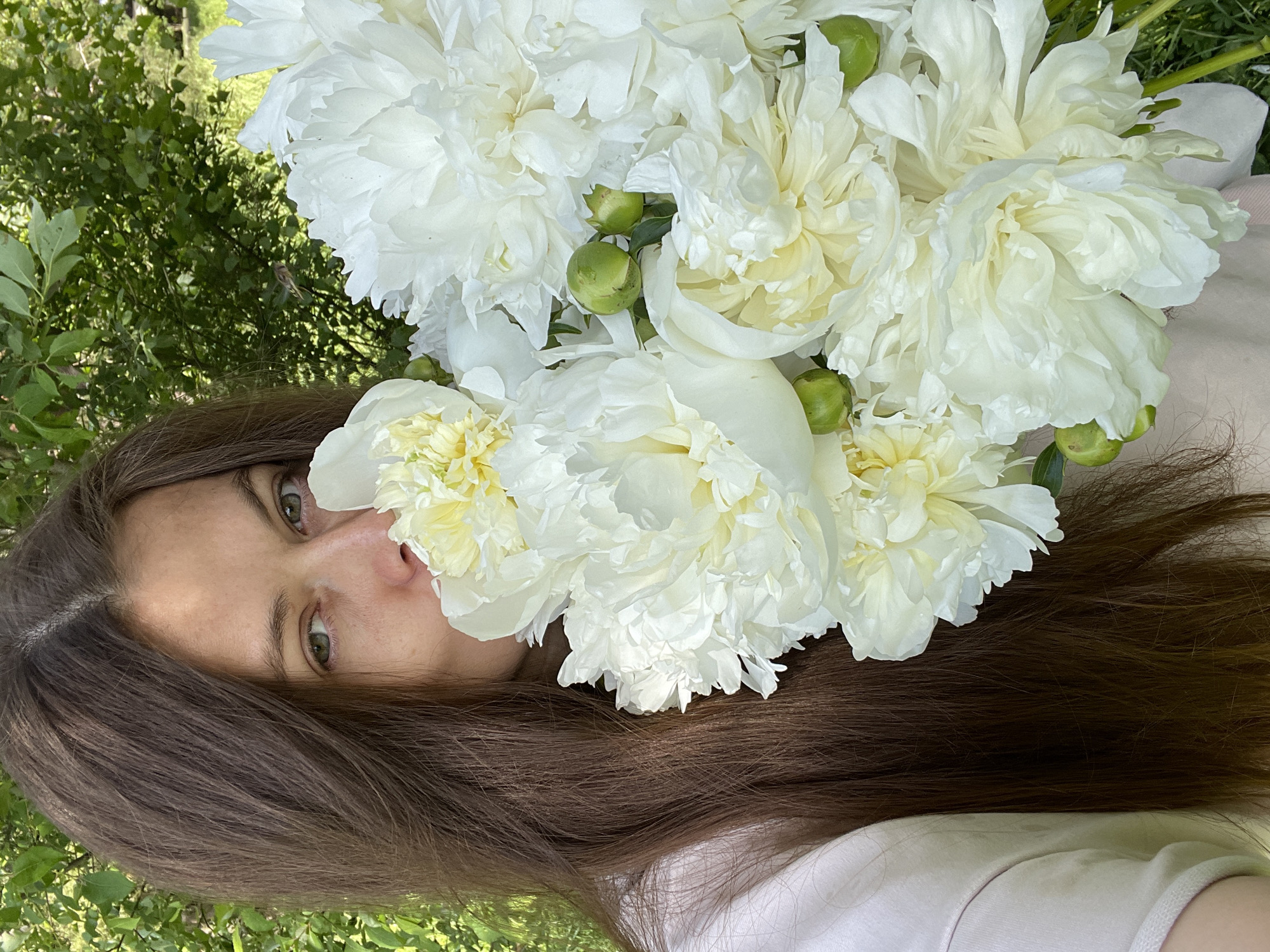 A woman with long, dark brown hair is holding a bouquet of white peonies in an outdoor setting. She is partially obscured by the flowers, with only her eyes and nose visible. The background is filled with green foliage, suggesting a garden or natural environment. The image appears to be taken in the city of Zarechny, Russia, though no specific landmarks are visible to confirm this. The lighting is natural and appears to be daytime. The woman is wearing a light-colored garment, possibly a t-shirt or blouse. The peonies are fully bloomed and appear fresh, with some unopened buds also present.