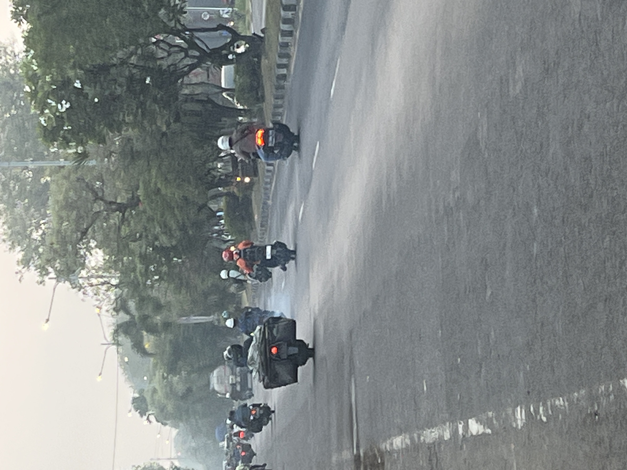 The image captures a road in Ngaban, Indonesia, with a procession of motorcycles moving along it. The atmosphere appears overcast or misty, with streetlights visible in the background. The road is asphalt, marked with white lines. On the right side of the road, there is a sidewalk or divider with a black and white striped border, followed by a line of trees. On the left side of the road, the surface appears wet, suggesting recent rain.
Several motorcycles are visible, some carrying riders and some with luggage. One motorcycle in the foreground is carrying a large, dark cargo box. Another motorcycle further back has a rider wearing an orange jacket and a white helmet. In the distance, more motorcycles can be seen, along with what appears to be a large truck. The trees in the background are lush and green.