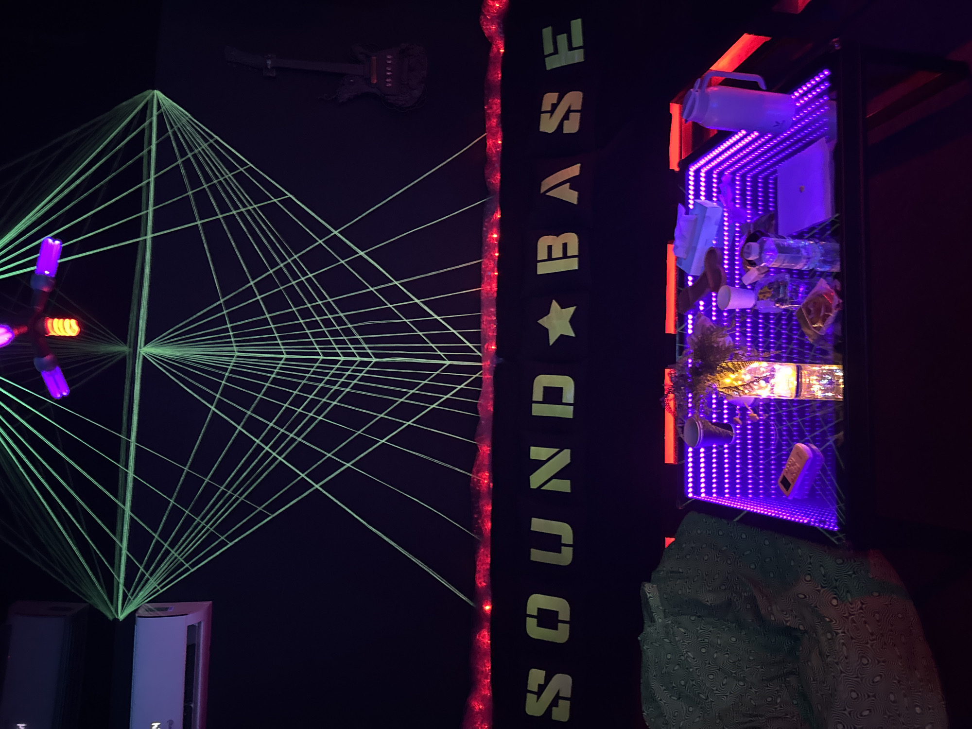 The image shows an indoor setting, likely an entertainment venue, featuring several distinct visual elements. On the left, a geometric structure comprised of green laser beams creates a network of intersecting lines. Above this laser setup, a decorative guitar is mounted on the wall. The background is predominantly black.
Next to this laser setup, the word "SOUNDBASE" is written vertically with a star symbol. The text appears to be illuminated with a pale green light. A string of red lights runs along the side of the word.
To the right, there is an illuminated shelf or display case filled with various items, including a white water cooler container, a laptop, bottles, a coffee cup, and other miscellaneous objects. The shelf is bordered with bright purple lights, and the entire structure is framed with red light strips.
A person wearing a patterned shirt is partially visible at the bottom of the image, near the display case. The location and specific city are not identifiable based on the visual information provided.