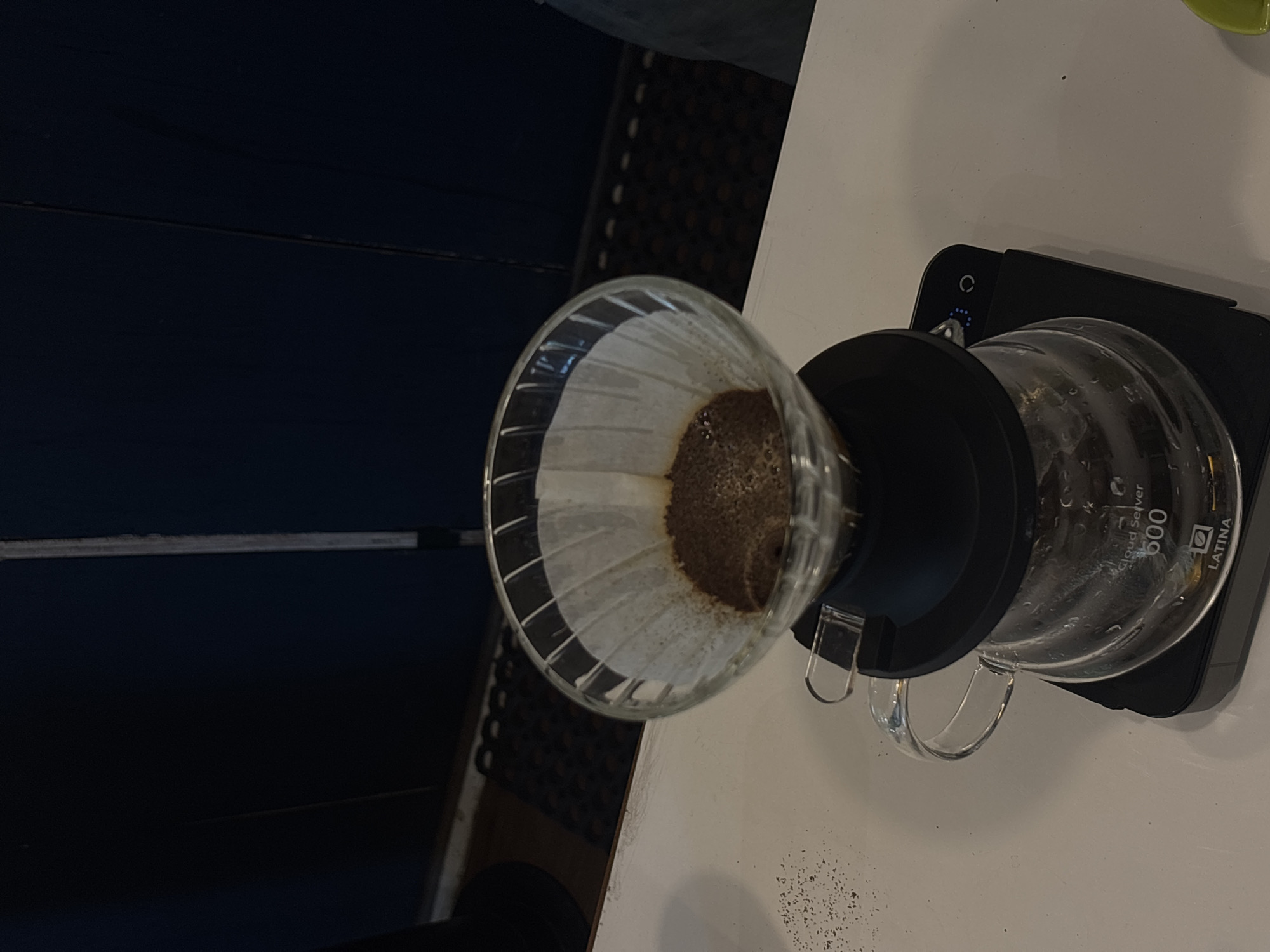 An overhead shot captures a pour-over coffee setup on a white countertop. The setup includes a glass dripper with a paper filter containing coffee grounds, resting on a glass server with measurement markings and the "LATINA 600" logo. The server sits on a black digital scale with blue indicator lights. In the background, dark blue panels are visible, and a black mat with a repeating pattern is on the floor. The image does not provide any information to indicate a location in Semarang, Indonesia.
