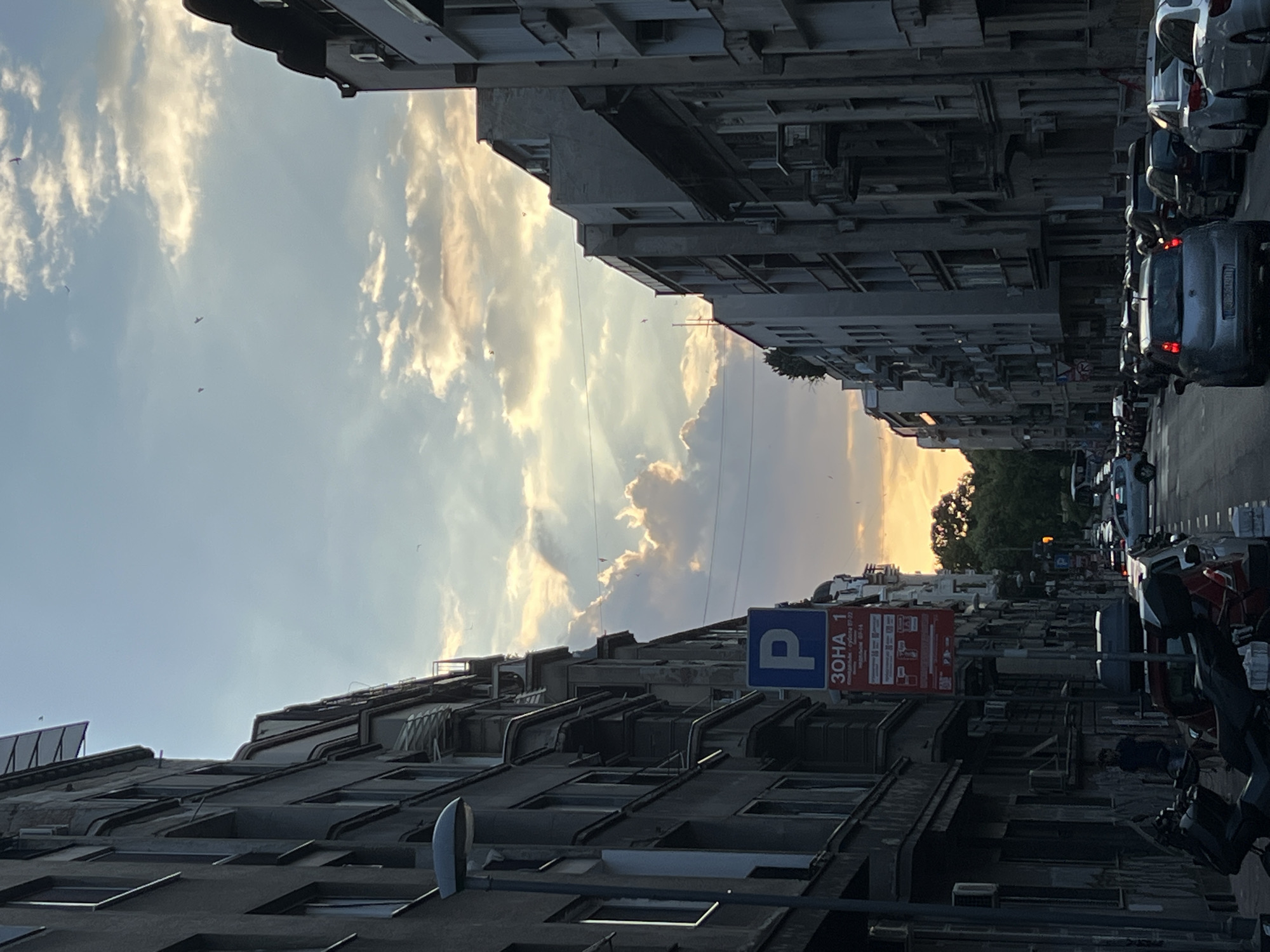 The image captures a street scene in Belgrade, Serbia, likely during the late afternoon or early evening. The sky is partly cloudy with the sun setting behind a band of trees in the distance, casting warm, orange light on the clouds. Tall, multi-story buildings with balconies line both sides of the street, creating a canyon effect. The architecture appears to be of an older European style.
In the foreground, on the left side of the street, there is a lamppost and a blue parking sign with Cyrillic text that reads "ЗОНА 1" (Zone 1). Below this, there is additional signage related to parking regulations. A motorcycle is parked to the left of the sign. Further down the street, a person in a dark shirt is standing near the buildings.
The street itself is lined with parked cars on both sides, with a traffic lane in the center. Several cars are visible, including a silver hatchback in the middle ground, facing away from the viewer. Traffic appears to be moving slowly or stopped in the distance. There are also smaller birds, or possibly kites, visible in the sky. The overall impression is of a typical urban street in a European city with a mixture of residential and commercial buildings, with the natural light of the sunset adding a dramatic element to the scene.