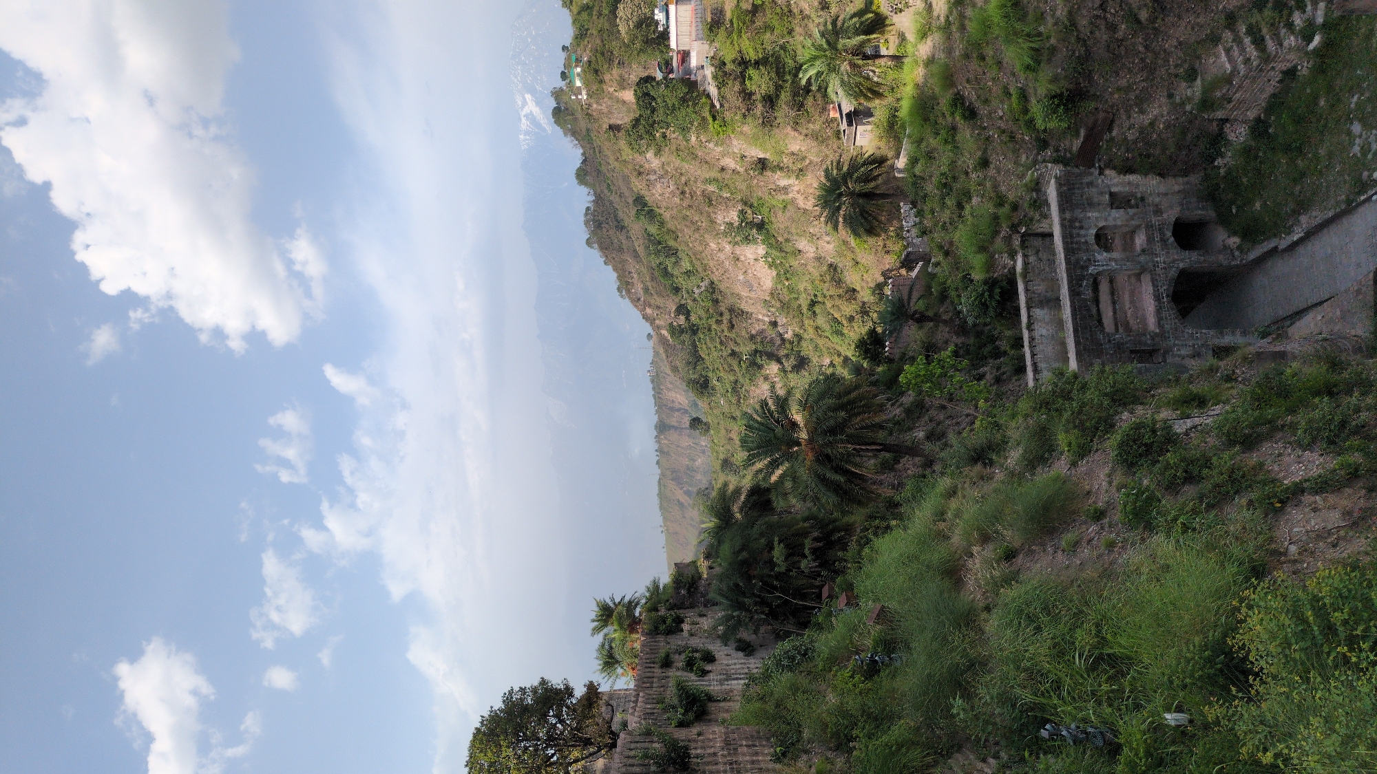 The image shows a landscape with a combination of natural and man-made elements. The sky is visible with scattered white clouds. A mountainous region occupies the central part of the image, densely covered with trees and vegetation. There is a building or structure along the ridge, further adding to the landscape's complexity. In the lower right corner is a ruined stone structure with arches and a walkway.
This is likely located in Kaswara, India, given the context provided. No people or animals are directly visible. The image captures the intertwining of nature and historical architecture.