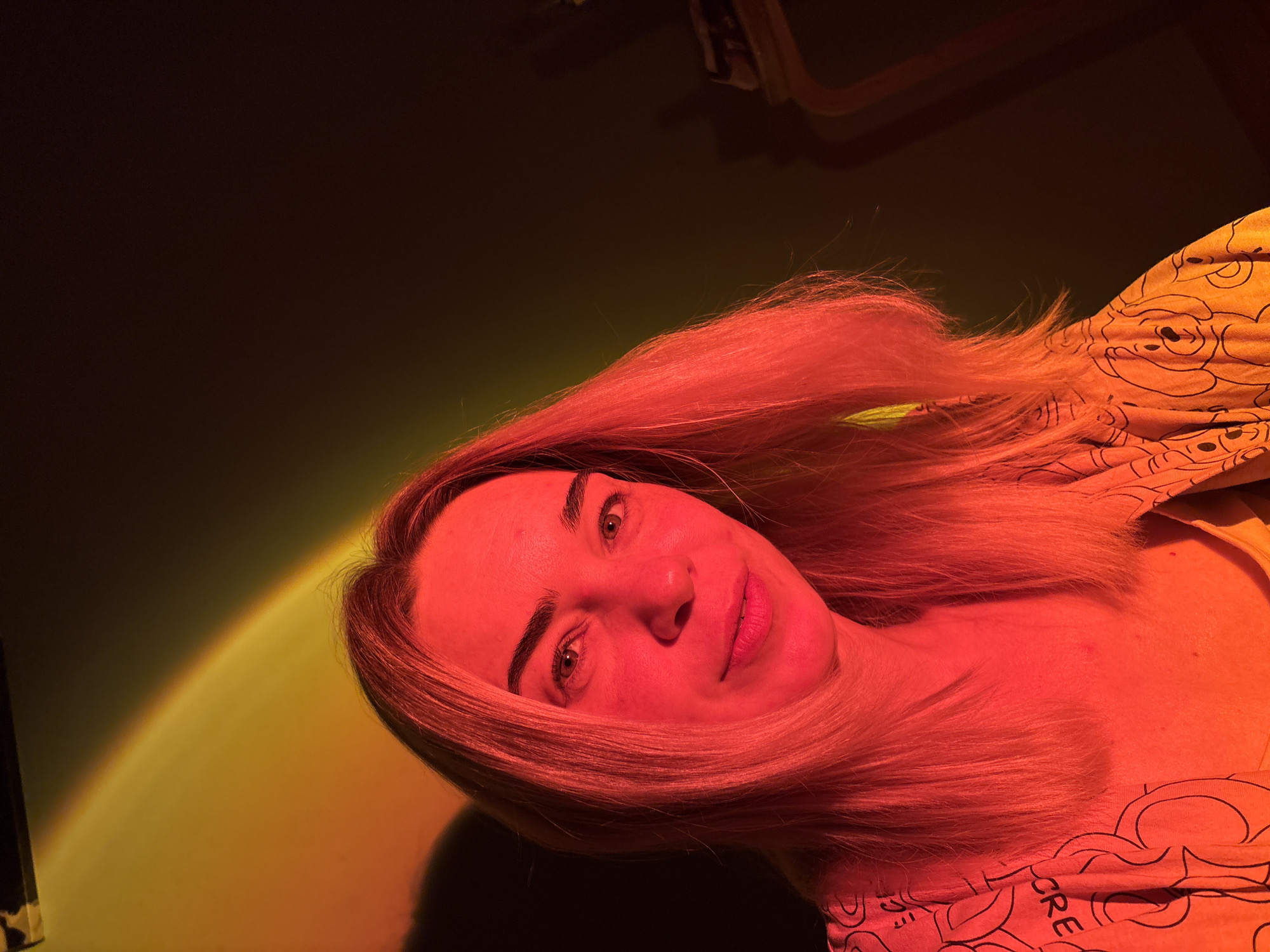 A medium shot shows a woman from the shoulders up, looking directly at the camera. She has blonde hair and her face is illuminated by red and yellow lighting, giving it a warm glow. She is wearing an orange top with a pattern of cartoon bears. The background is dark, with a blurred reflection of the lighting on the left side of the image. There are no discernible objects or landmarks that would indicate a specific location in Sinyavino, Russia. The image is a self-portrait.