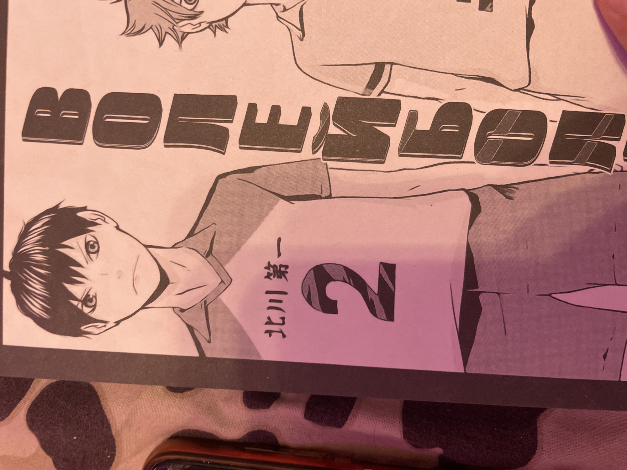The image is a close-up photograph of a manga panel featuring two male characters in athletic uniforms. The character on the left is wearing a collared shirt with the number "2" prominently displayed on the chest, along with Japanese text above it. The character on the right, only partially visible, appears to be wearing a similar uniform. The background is a stylized, abstract pattern with dark spots. Large, bold, dark letters are positioned vertically to the right of the characters, suggesting a title or inscription. The lighting in the image is warm, casting a pinkish hue over the scene. The photograph is taken at a slight angle, and a portion of a mobile phone and a patterned fabric are visible in the lower left corner of the image. There is no identifiable information within the image that suggests a location in Irinovka, Russia.