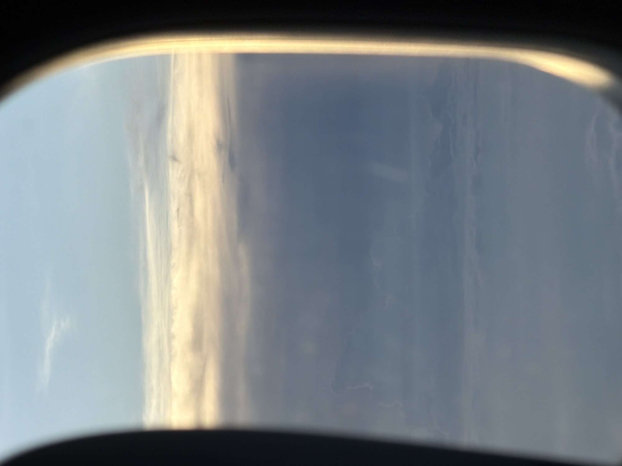 The image is an aerial view from an airplane window, looking out at a sky filled with clouds. The sky transitions from a pale blue at the top to a hazy, lighter blue towards the bottom. Several layers of clouds are visible, with the upper layers appearing wispy and light, catching the sunlight and appearing golden or white. Below these, denser, darker blue clouds form the bulk of the view. The edge of the airplane window frame is visible on the left and right sides of the image, with the right side showing a strong, bright reflection of sunlight. The image does not provide any details about the city of Leh, India.