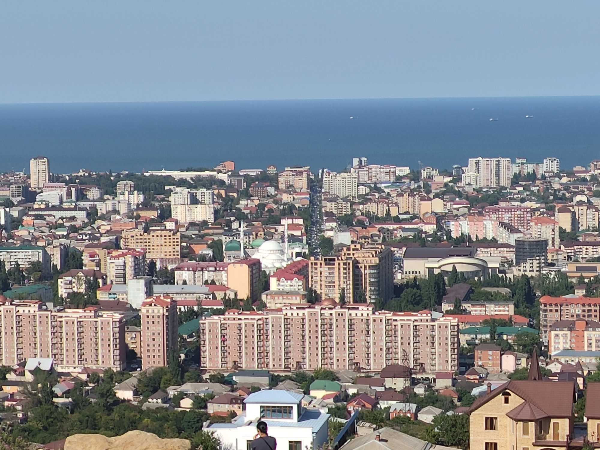 The image shows a panoramic view of the city of Makhachkala, Russia, with the Caspian Sea in the background. The foreground features a rocky outcrop, while the midground is densely populated with apartment buildings, ranging in height from a few stories to taller, multi-story structures with visible balconies. Several large, light-pink residential complexes dominate the central part of the frame. Smaller houses with pitched roofs are scattered among the larger buildings. Green trees are interspersed throughout the urban landscape. In the distance, the blue expanse of the Caspian Sea meets a clear sky, with a few ships visible on the water. One person, partially visible from the back, stands on a rooftop in the lower right quadrant of the image. The overall scene depicts a sunlit urban environment bordering the sea.