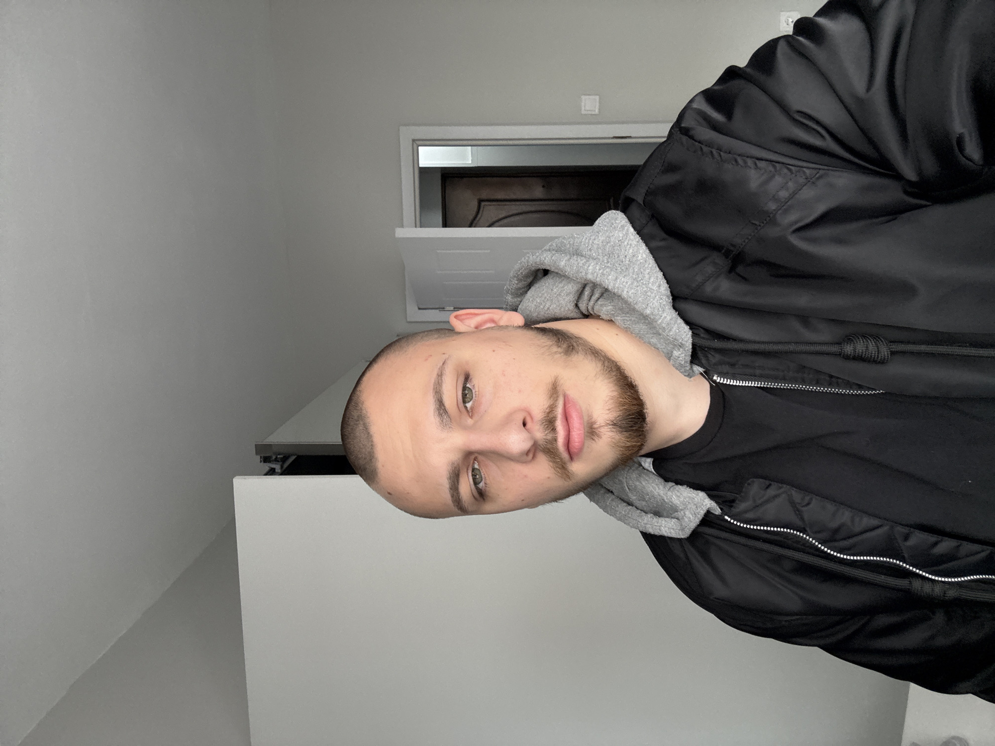 A man with close-cropped hair, a beard, and wearing a black jacket over a gray hooded sweatshirt is indoors. He is facing the camera. The background includes white walls, a door, and a partially open attic access panel. The location is not identifiable as being in Stara Zagora, Bulgaria from the image alone.