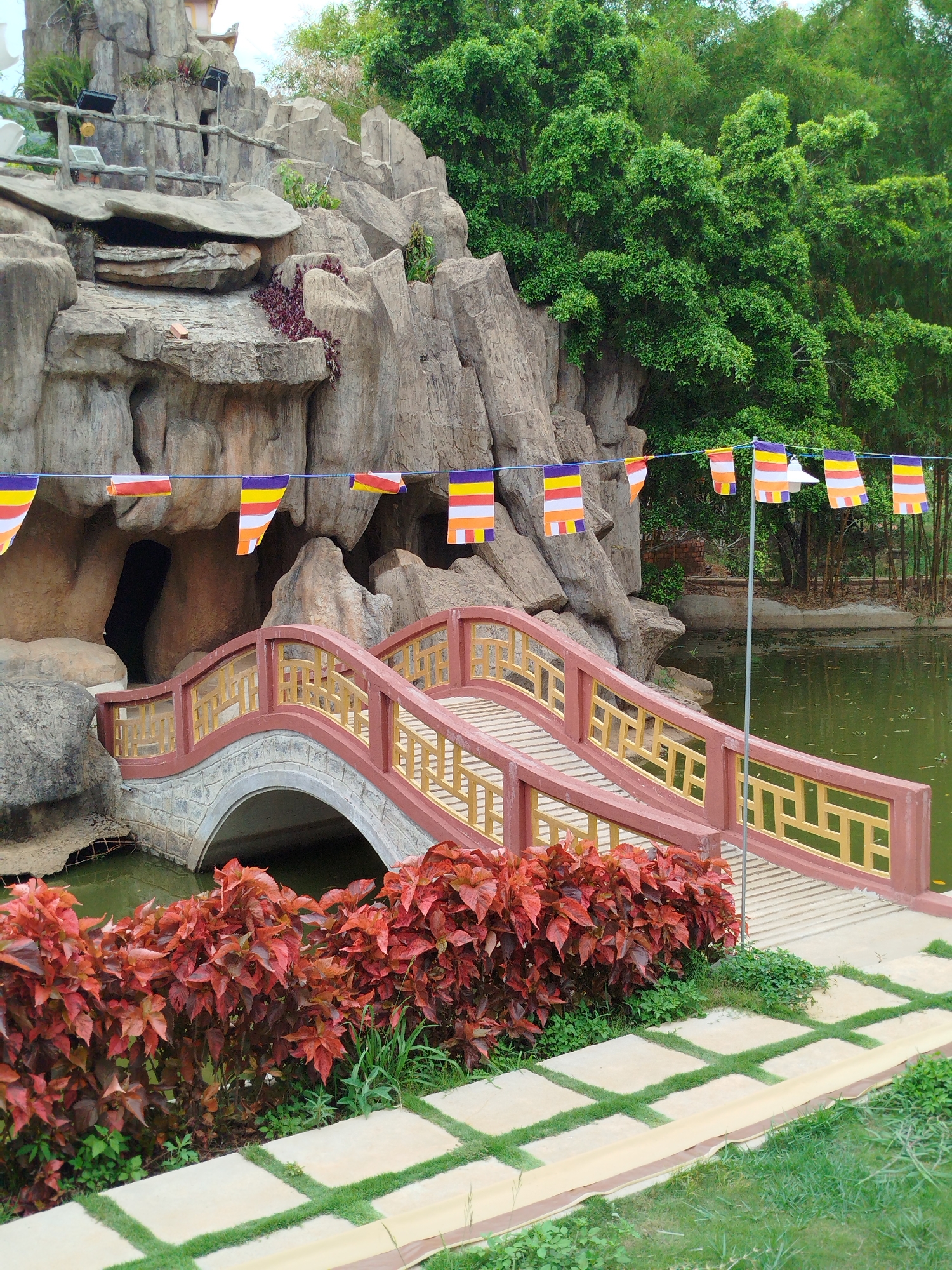 This tranquil daytime photograph captures a scenic garden or temple ground in Da Lat, Vietnam. A prominent feature is an elegantly designed pedestrian bridge with an arched base made of stone or concrete, and decorative red-brown railings with yellow, patterned balustrades. The bridge spans over a calm, dark green pond or stream, reflecting the surrounding greenery.
Above the bridge, a string of colorful rectangular Buddhist prayer flags, displaying horizontal stripes of blue, yellow, red, white, and orange, are strung across the scene, adding a spiritual touch. These flags are supported by a thin metal pole on the right side of the bridge.
The background is dominated by impressive, large greyish-brown rock formations, which appear to be either natural grottoes or artfully constructed rockeries, some featuring small cave-like openings. Small red-leafed plants grow in crevices of these rocks, and spotlights are visible, suggesting the area is illuminated at night.
Lush vegetation surrounds the scene; vibrant red-leafed bushes line the foreground pathway, which is made of light-colored square paving stones with grass growing in the gaps. Beyond the rock formations, tall, dense green trees and bamboo create a verdant backdrop. The overall atmosphere is peaceful and serene, with no people visible, suggesting a quiet place for reflection or a leisurely stroll. No visible text is present in the image.