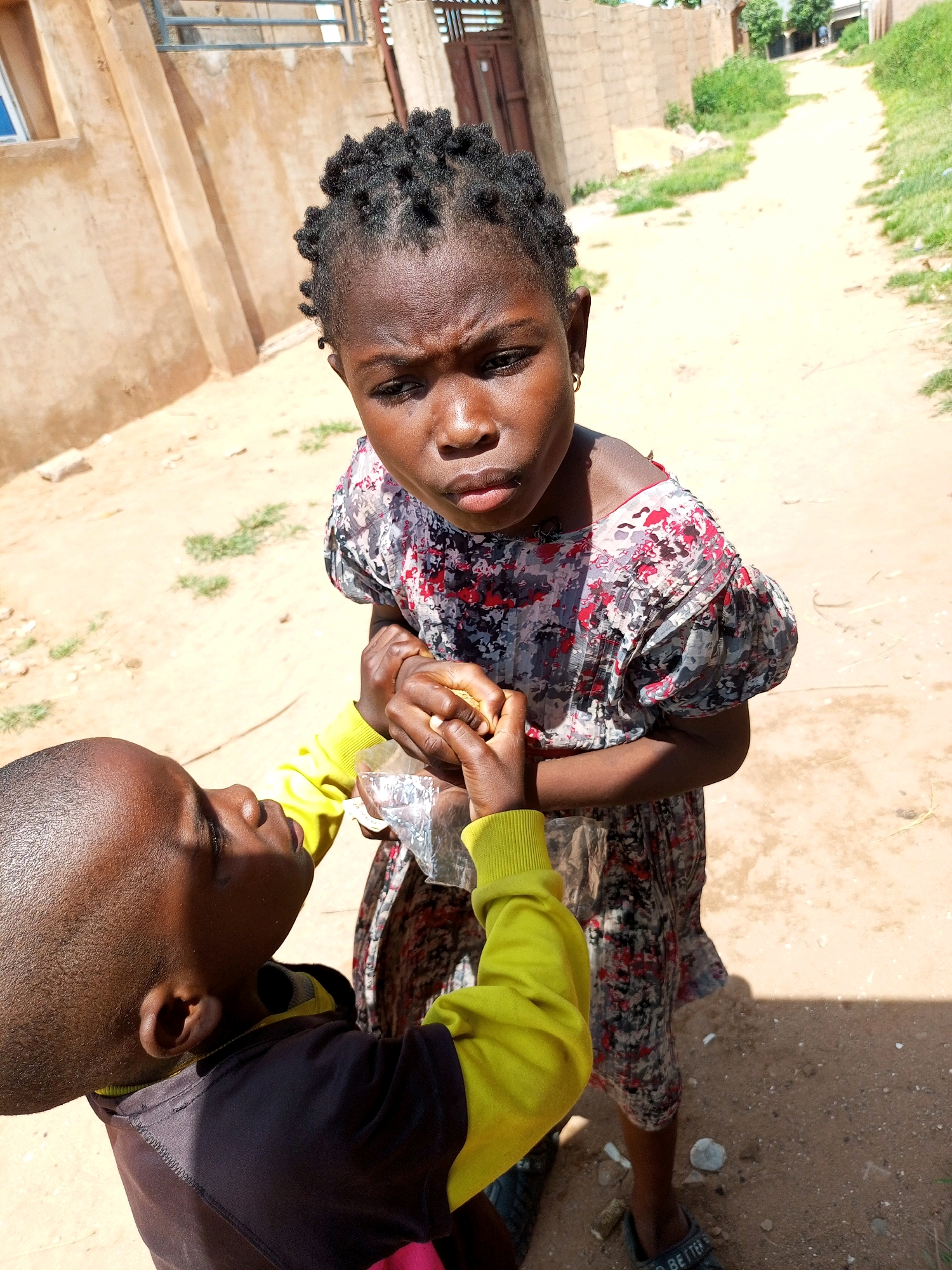 A high-angle, medium shot shows two young children interacting outdoors on a dirt path. The child on the right, a girl with dark skin and braided hair, wears a floral-patterned dress with red and grey designs and a gold hoop earring in her left ear. She is looking down with a pensive expression, her left hand extended towards the child on the left. The child on the left, a boy with dark skin and a shaved head, wears a dark long-sleeved shirt with yellow sleeves and a black hood. He is holding a clear plastic bag in his hands and looking up at the girl. His left hand is reaching out to the girl. Both children's hands are close together, suggesting an exchange or shared moment. The environment appears to be a rural or semi-urban area in Tempure, Nigeria, with a sandy dirt path stretching into the distance, lined by buildings with sandy-colored walls and some overgrown vegetation. The sunlight casts shadows on the ground, indicating it is daytime.