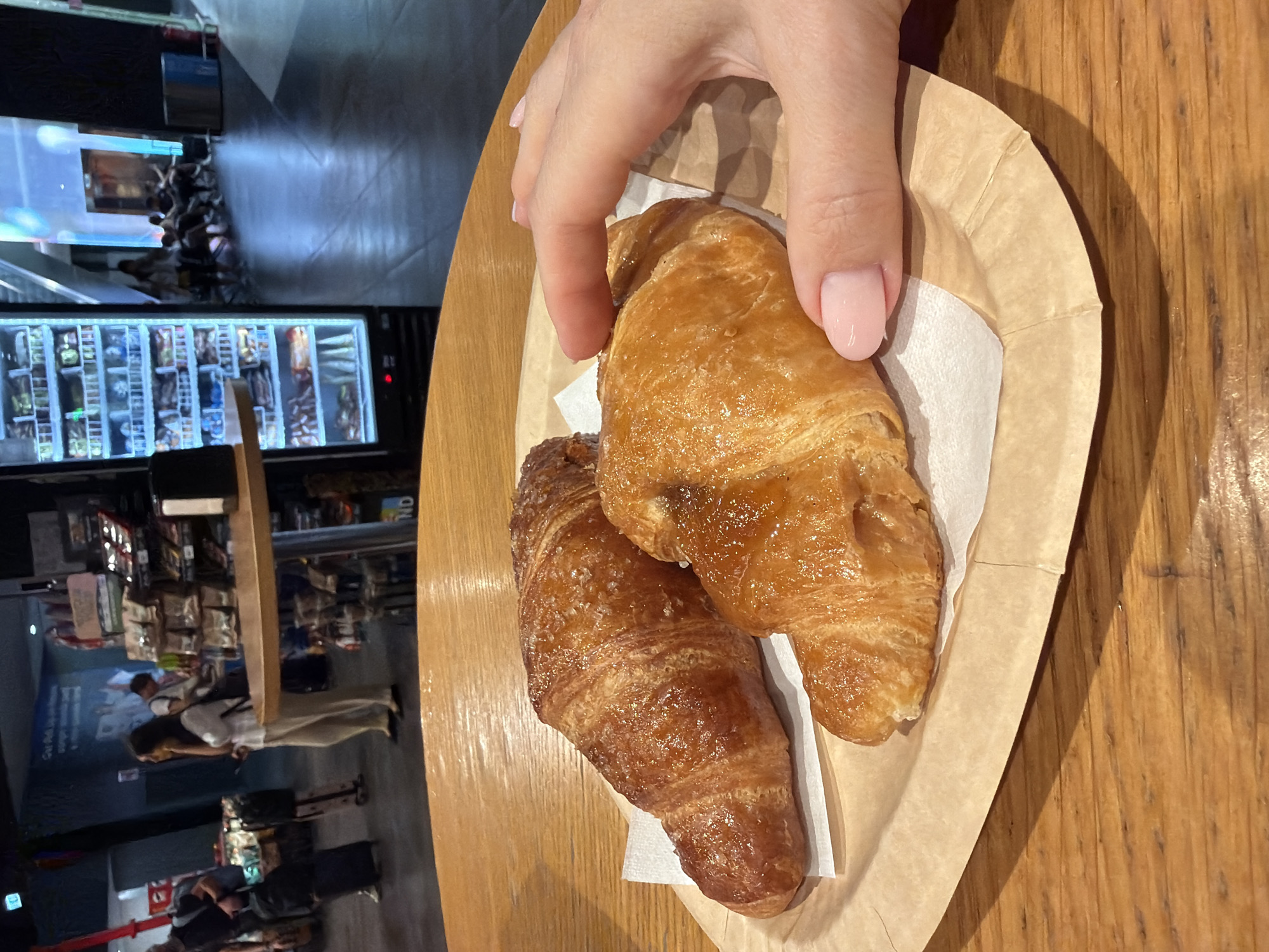 The image shows two croissants on a paper plate, placed on a wooden table. A woman's hand with pink nail polish is reaching for one of the croissants. In the background, there is a store with shelves stocked with food items and drinks. People can be seen in the background, some seated and some standing. The overall setting appears to be an airport or a similar transit hub, possibly Fiumicino Airport in Italy, given the context of the location. The lighting is a mix of ambient and artificial lights from the store displays.