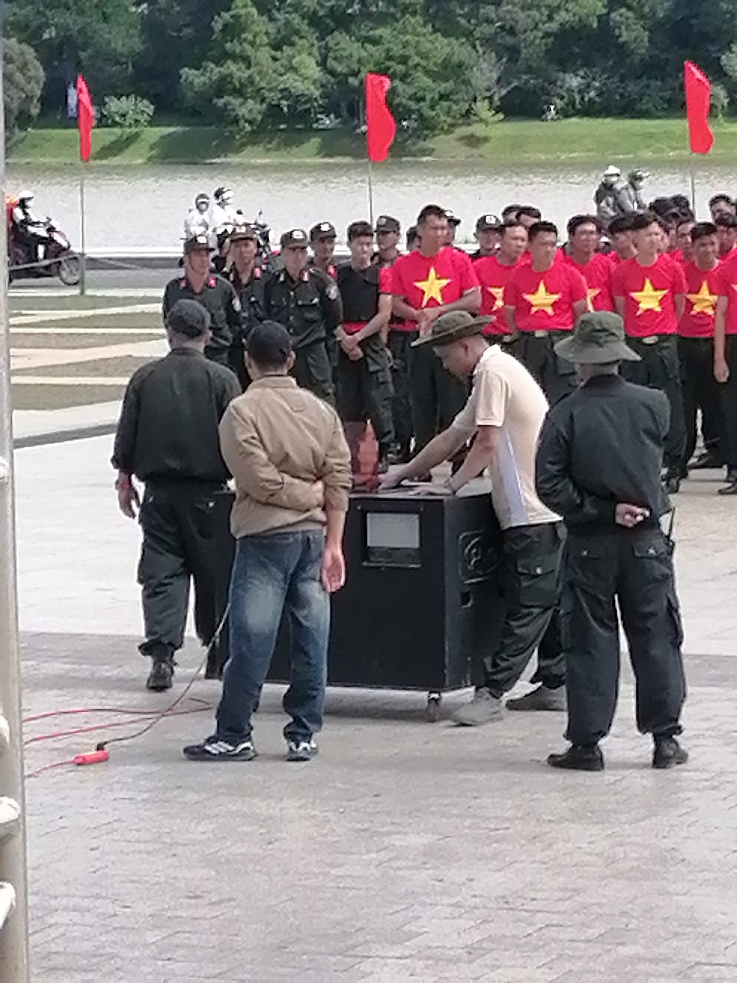 The image depicts a gathering of individuals, many in uniform, outdoors. The setting appears to be a public space, possibly near a body of water, with trees and greenery in the background. Several people are wearing red t-shirts with a yellow star, suggesting a national or organizational affiliation. Some individuals are in military or police attire, with caps and dark uniforms.
A prominent black structure, possibly a sound system or equipment case, is in the center foreground. One person in a light-colored shirt and a hat is leaning over this structure, appearing to interact with it. Another person in a dark uniform stands behind it, with hands clasped behind their back. To the left, two individuals in dark uniforms are walking away from the camera, one with a red cord attached to their leg. In the middle ground, a line of people in red t-shirts with stars are standing, and further back, more uniformed personnel are visible, along with some motorcycles. Red flags are prominently displayed on poles in the background.
The presence of red flags, the style of the uniforms, and the general ambiance suggest a formal event or ceremony. Given the visual cues such as the attire of the individuals and the national symbols, it is highly probable that this scene is located in Da Lat, Vietnam, a city known for its historical significance and cultural events. The overall impression is of an organized assembly, possibly a public address, ceremony, or a demonstration.