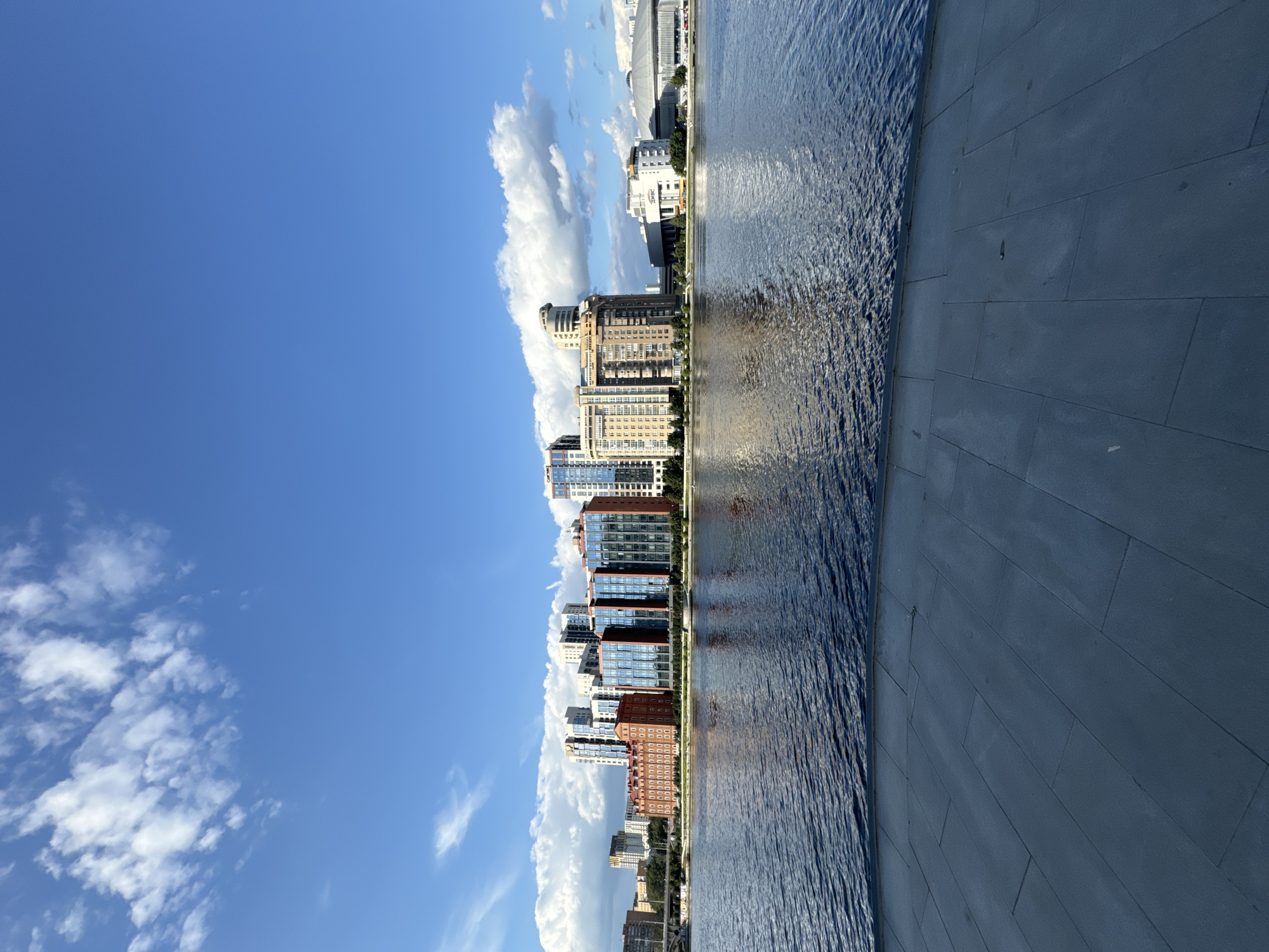 This is a vertical, eye-level, wide shot of a cityscape with a body of water in the foreground. The scene depicts the waterfront of a city with a clear blue sky and scattered white clouds. A paved promenade in a dark gray tone runs across the bottom of the frame, with large rectangular tiles laid out in a linear pattern. Beyond the promenade, a wide, dark blue body of water stretches horizontally across the middle of the image, reflecting the sky and buildings.
On the opposite bank of the water, a row of modern buildings forms the city skyline. These buildings vary in height and architectural style, with some featuring glass facades and contemporary designs, while others appear to be older brick structures. The buildings are illuminated by natural daylight, and their reflections are visible on the water's surface. The overall impression is one of an urban waterfront area on a bright, sunny day.