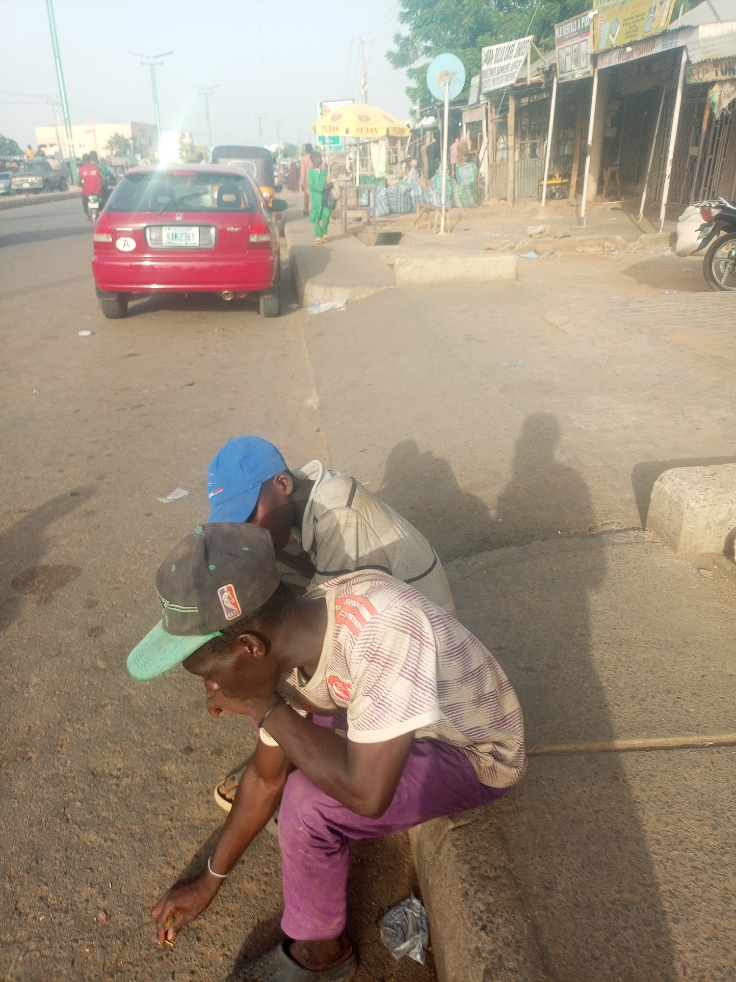 Two males are seated on a curb next to a street in Tempure, Nigeria. The first male, closer to the camera, wears a black and green NBA cap, a white striped shirt, and purple pants. He is looking down and appears to be picking something up off the street. The second male, sitting behind him, wears a blue cap and a light-colored long-sleeved shirt. Behind them is a red car parked partially on the street and partially on the curb. Further back, there is a street with pedestrians and vehicles, as well as storefronts lining the sidewalk. The general environment suggests a bustling urban area with shops and traffic.