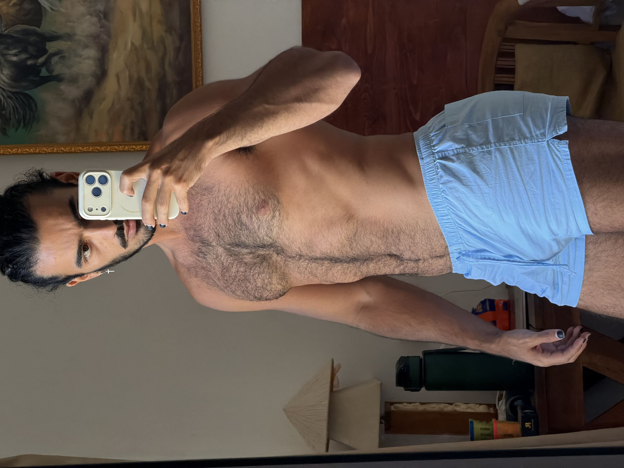 A man with dark hair pulled back into a bun and a beard is standing and taking a selfie in a room. He is wearing light blue boxer shorts and is shirtless. His left arm is raised, holding a white smartphone up to take the photo. He is wearing a silver cross earring in his left ear and has dark nail polish on his fingernails and toenails. The background shows a portion of a painting of horses on the wall, a lampshade, and a dark wooden dresser with items on it. The scene suggests the photo was taken indoors, likely in a bedroom, and not in a public space in North Kuta, Indonesia.