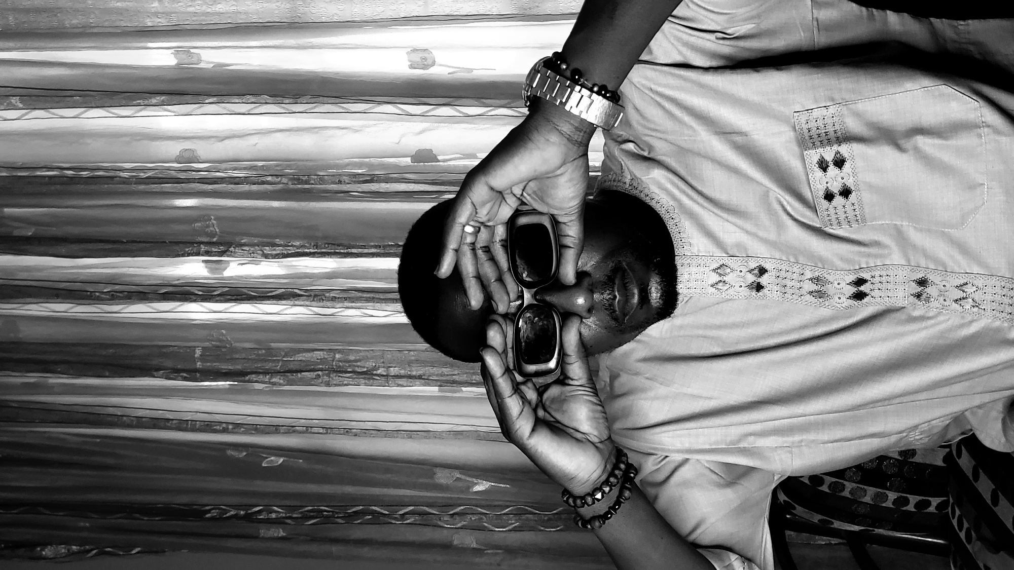 A medium shot of a Black African male in Sabon Tasha, Nigeria. The man is holding sunglasses to his face. He is wearing a light-colored, patterned shirt with short sleeves and a wristwatch and bracelets. He is positioned in front of what appears to be a curtain or decorative textile backdrop.