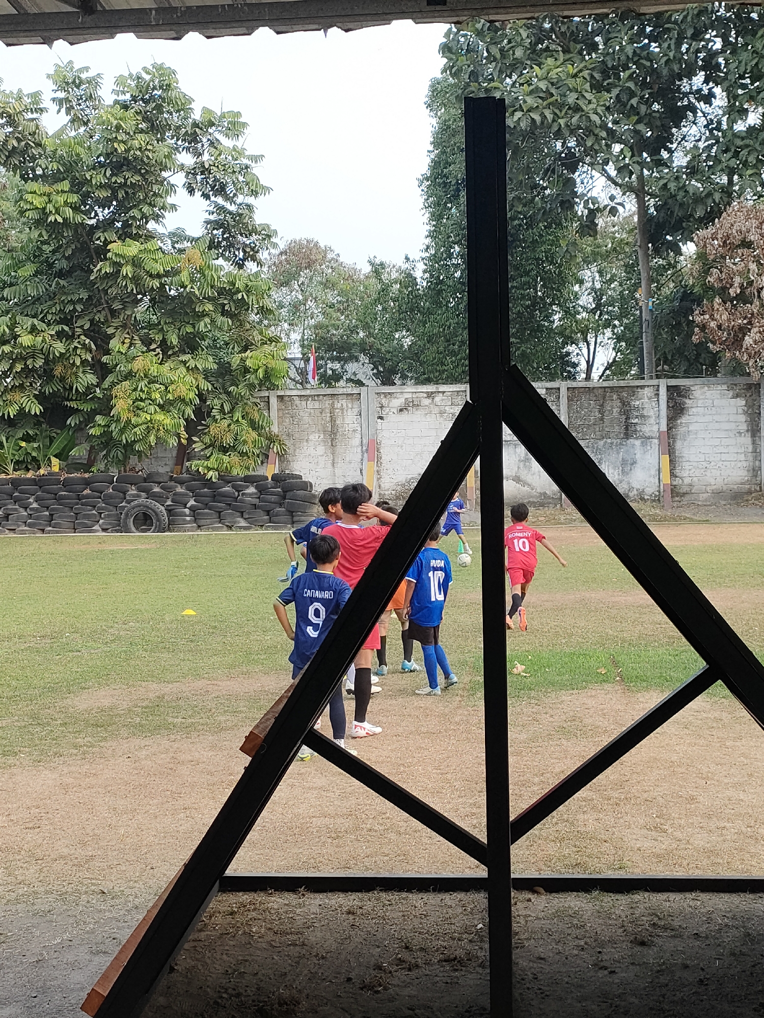 The image shows an outdoor soccer field in Banguntapan, Indonesia. Several young boys in soccer uniforms are visible on the field, some engaged in a game. One boy in a red jersey with "ROMENY 10" on the back is running. Another boy in a blue jersey with "CANAVARO 9" is standing. In the background, a white brick wall with some painted sections runs along the perimeter of the field, and a stack of old tires is piled up against it. Trees and foliage are visible behind the wall. A dark metal structure partially obstructs the view in the foreground. The ground is a mix of grass and dirt. The lighting suggests it is daytime.