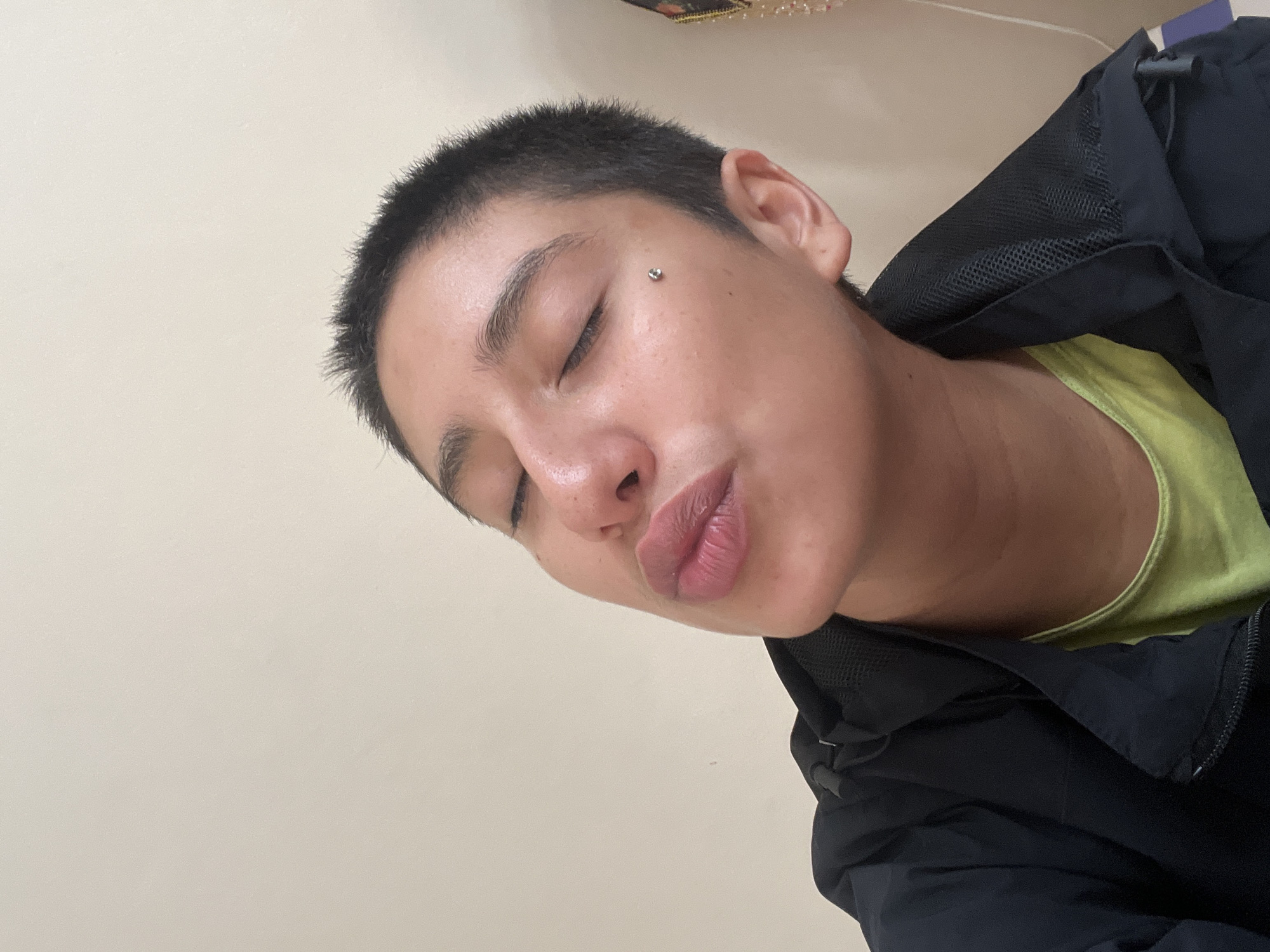 A person is lying down with their eyes closed and lips puckered, wearing a yellow shirt and a dark jacket. They have short, dark hair and a piercing on their cheek. The background is a plain wall. The location is not discernible from the image.